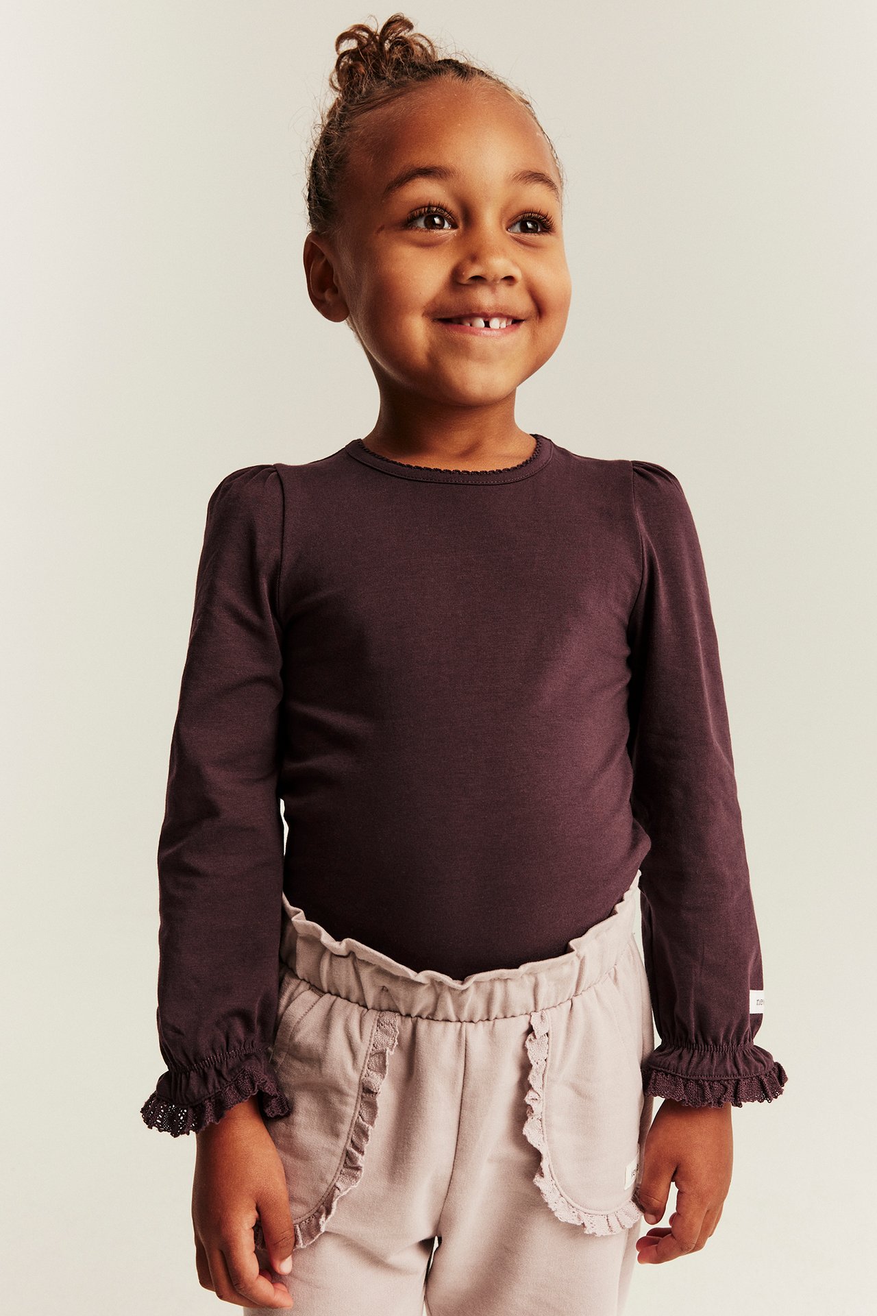 Front view, kid wears a dark brown long-sleeved top with ruffled cuffs and light beige ruffled pants.