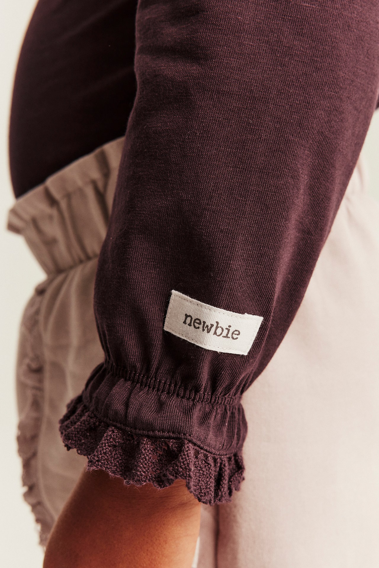 Nahaufnahme eines Babys in einem dunkelvioletten Langarmshirt mit Rüschenbündchen und beiger Hose.