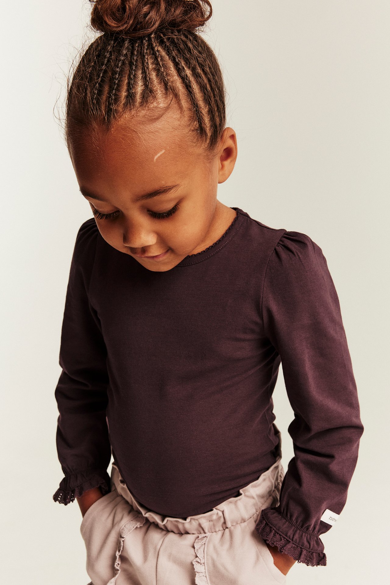 Front view, kid in dark purple long-sleeved top with ruffles and light beige pants.