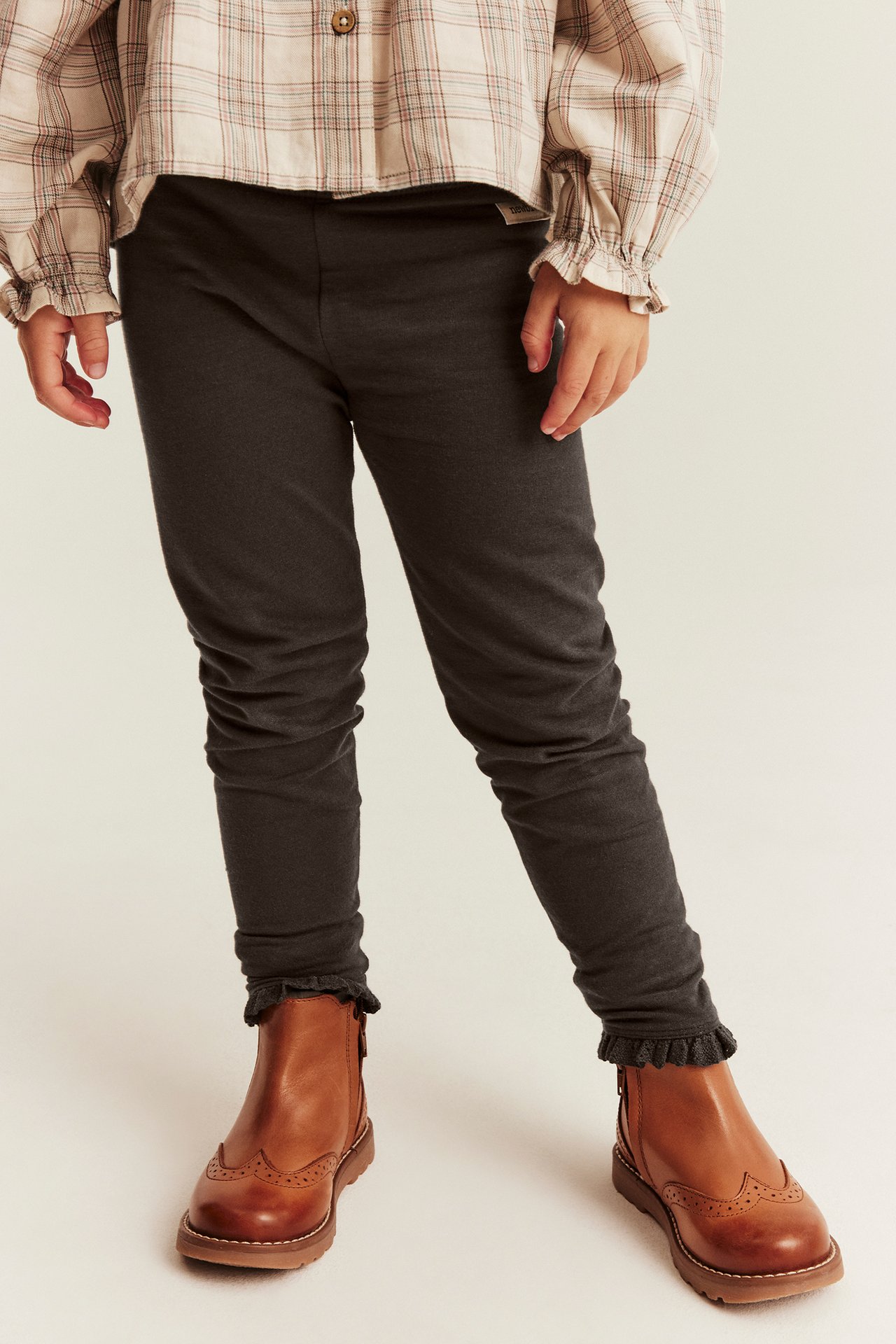 Vorderansicht eines Kindes in dunkelgrauen Leggings mit Rüschensaum, einem beigen Karohemd und braunen Brogues.