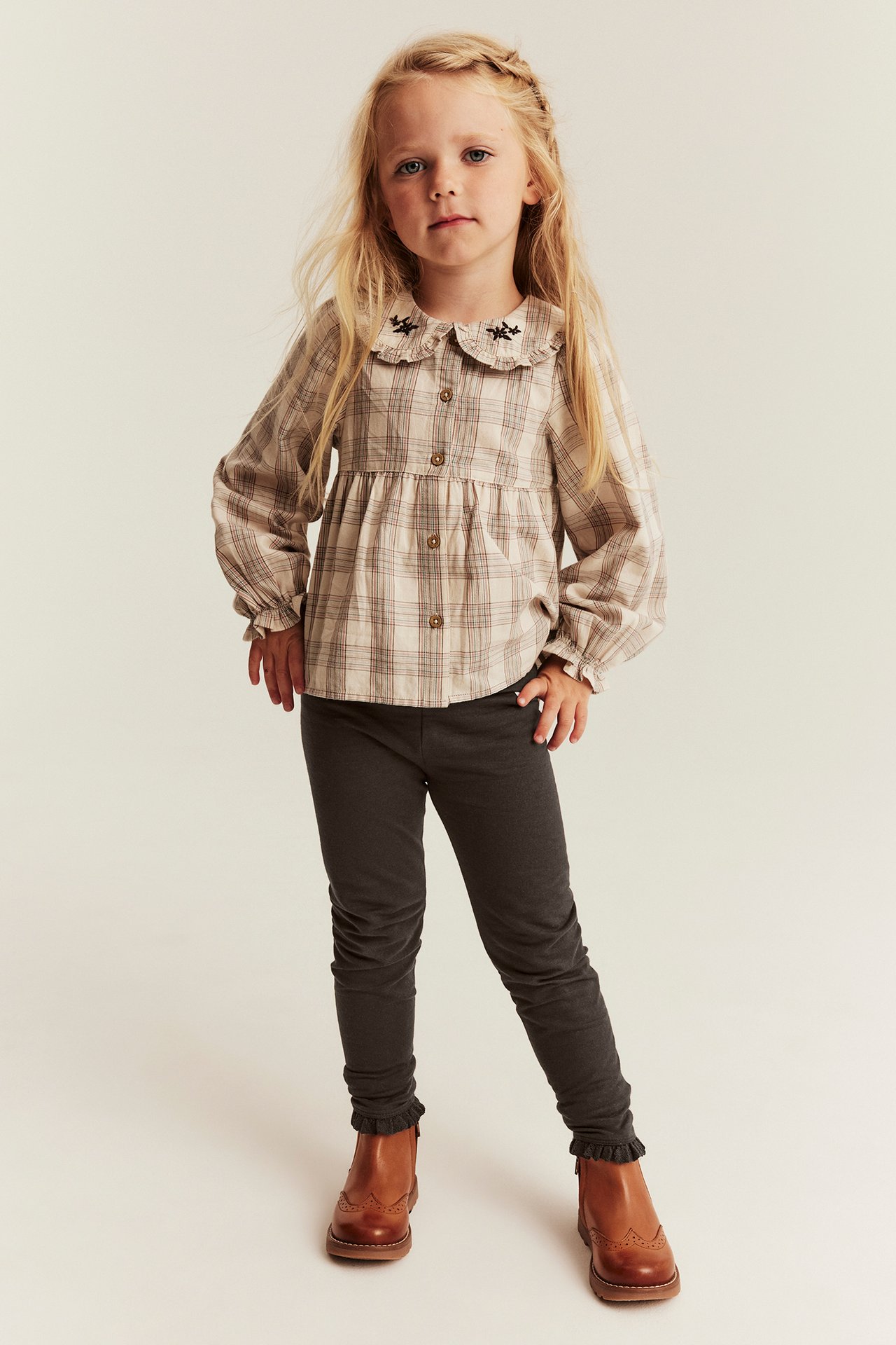 Mädchen, Vorderansicht: Beige karierte Peplum-Bluse, dunkelgraue Leggings, braune Brogue-Stiefeletten.