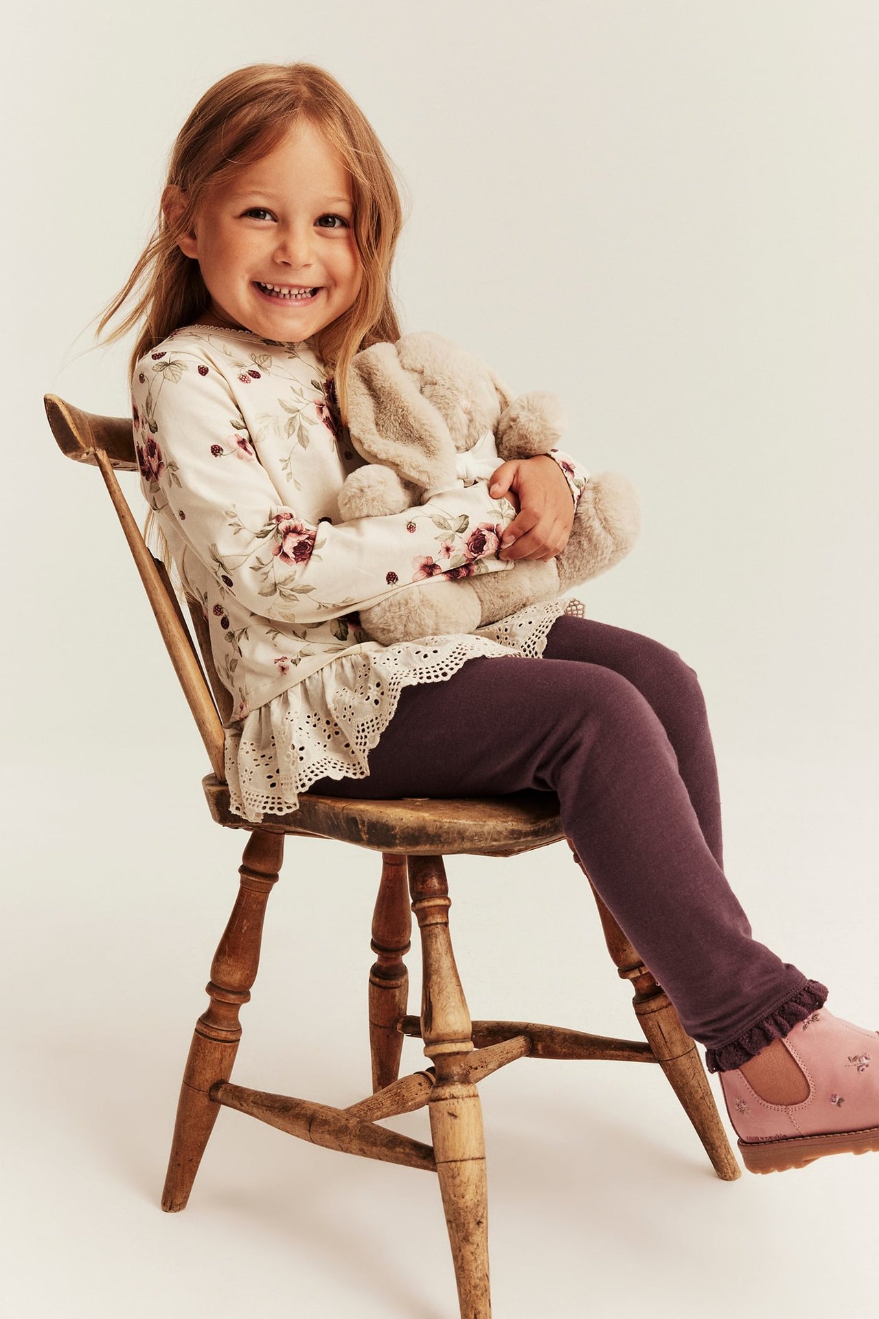 Kindertop mit Blumenmuster und Spitze, dunkelviolette Leggings, rosa bestickte Schuhe, hält Plüschkaninchen.