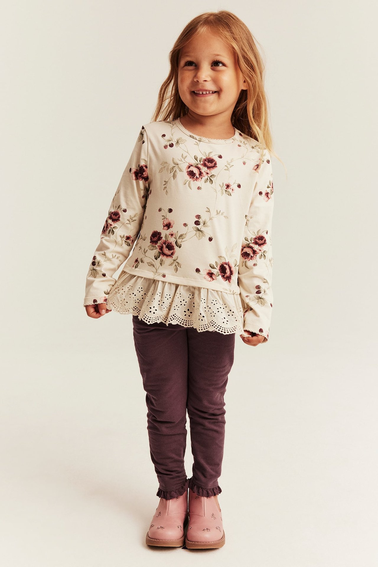 Vorderansicht Kind: blumiges Langarmshirt mit Lochstickerei, pflaumenfarbene Leggings, pinke Stiefel.