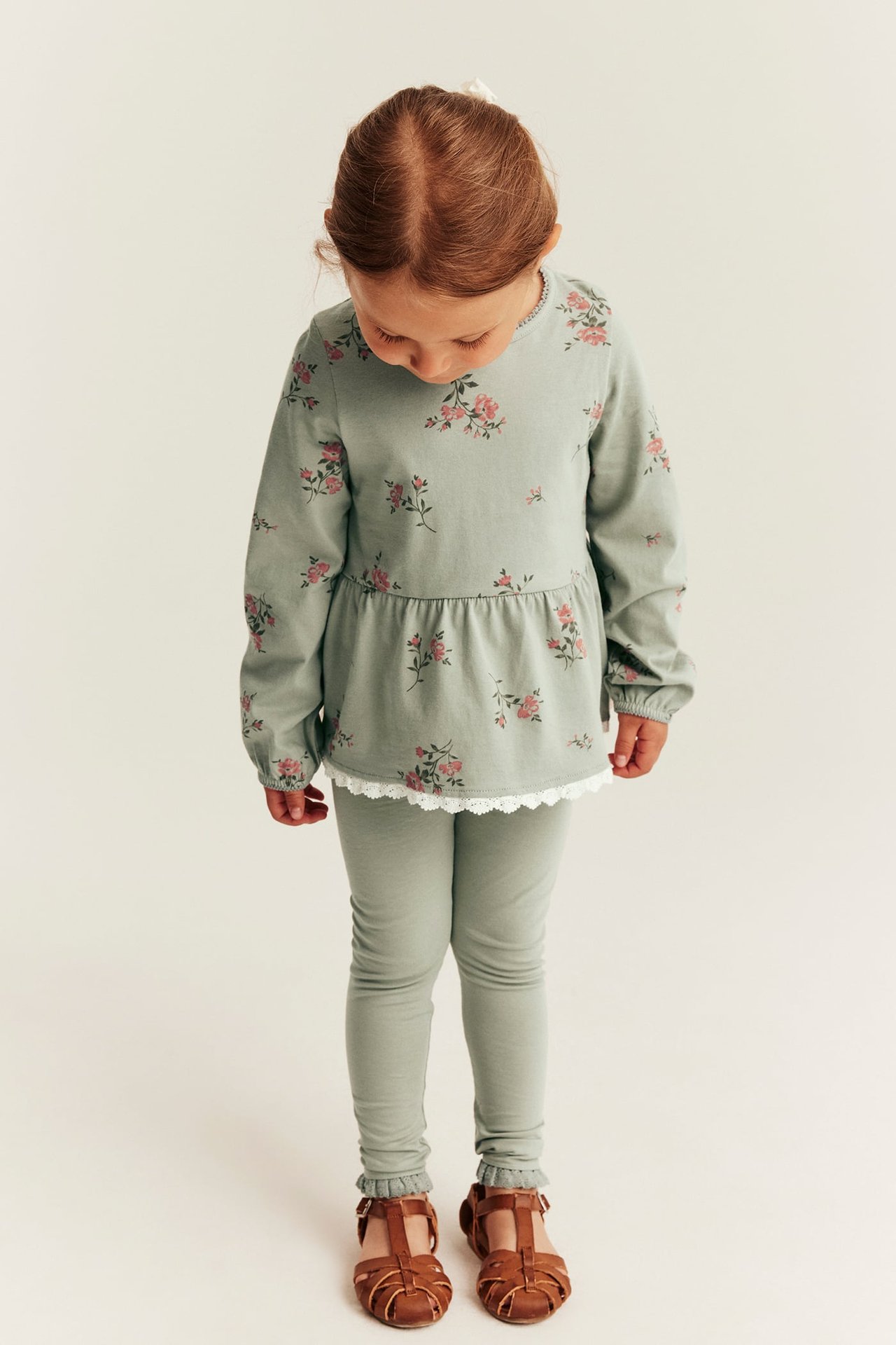Vorderansicht: Kind in grünen Leggings mit Spitze, geblümtem Peplum-Top und braunen Sandalen.