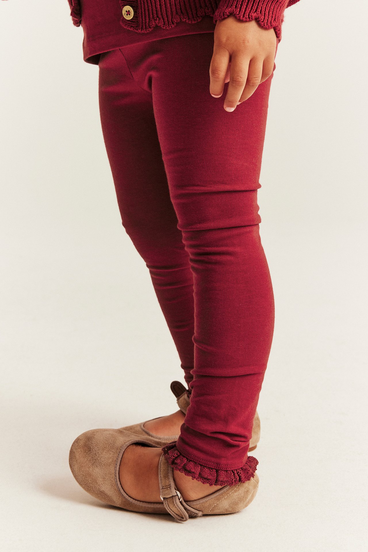 Seitenansicht eines Kindes in burgunderfarbenen Rüschen-Leggings, passendem Cardigan und beigen Ballerinas.