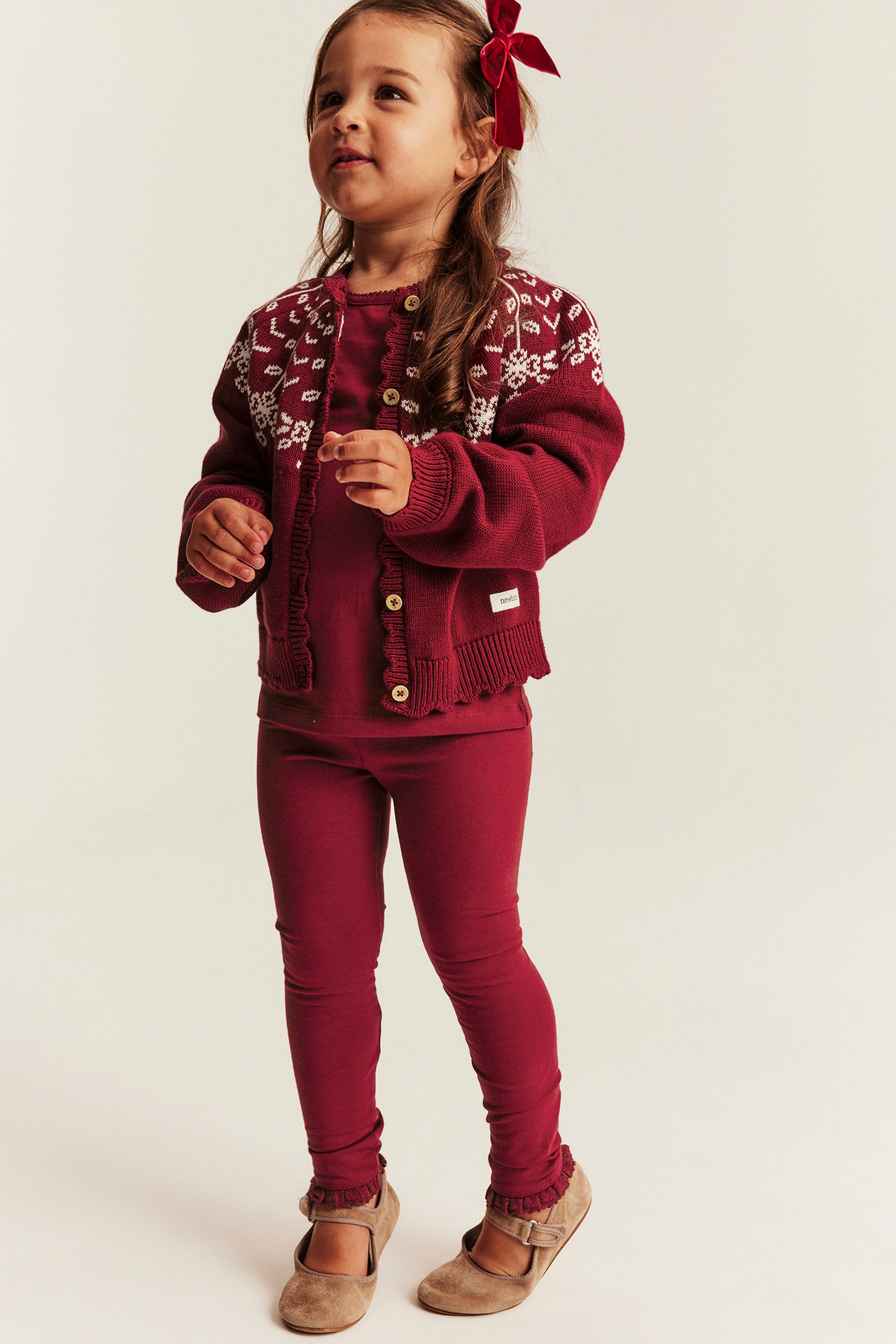 Vorderansicht: Kind in roter gemusterter Strickjacke, rotem Top, roten Leggings und beigen Spangenschuhen.