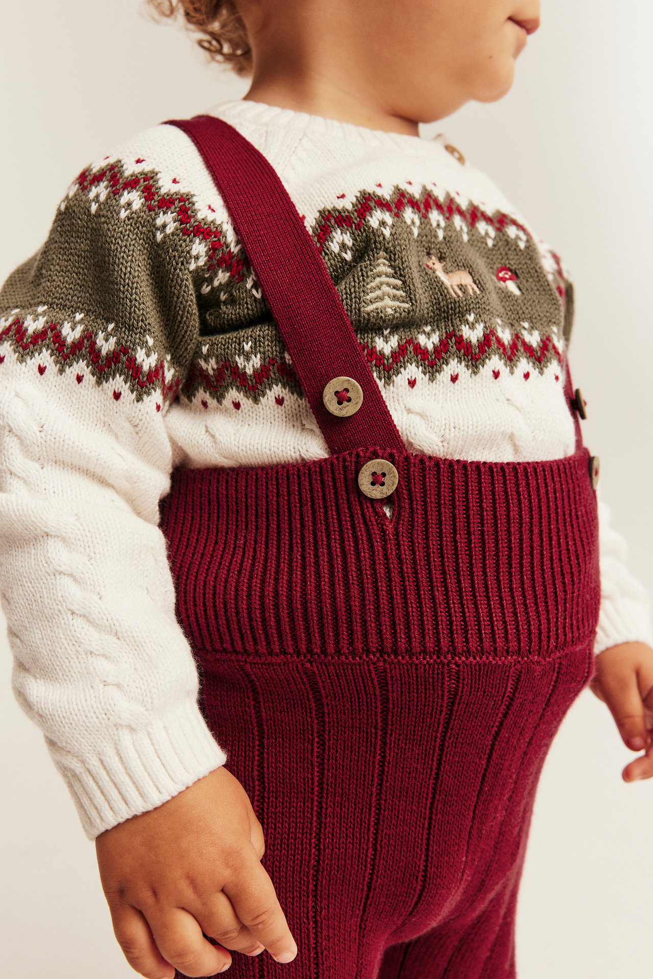 Nahaufnahme von Baby in burgunderfarbenen Rippstrick-Latzhosen über weißem Fair-Isle-Pullover.