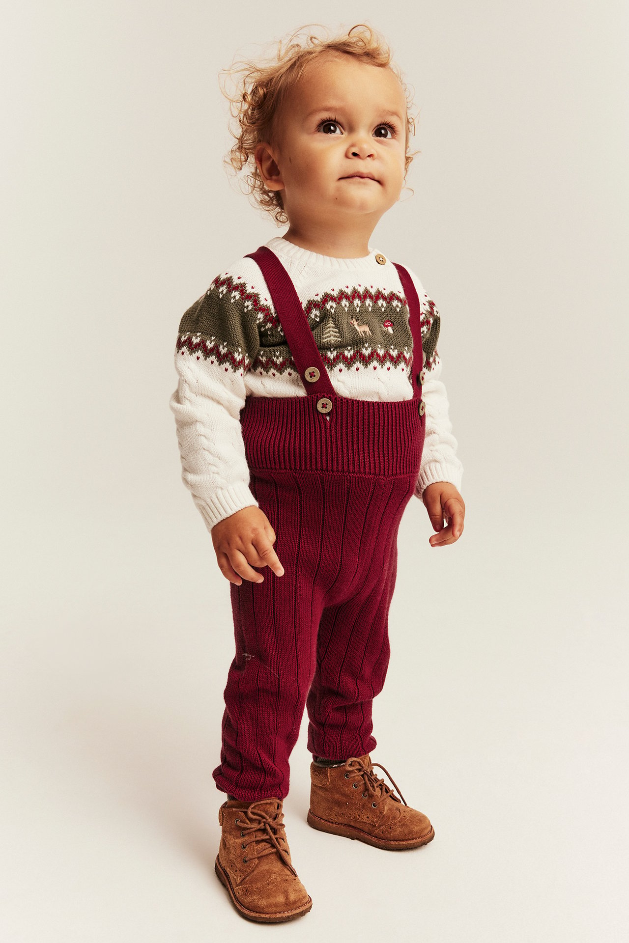 Vorderansicht von Baby in roter Stricklatzhose, weißem gemustertem Pullover und braunen Stiefeln.