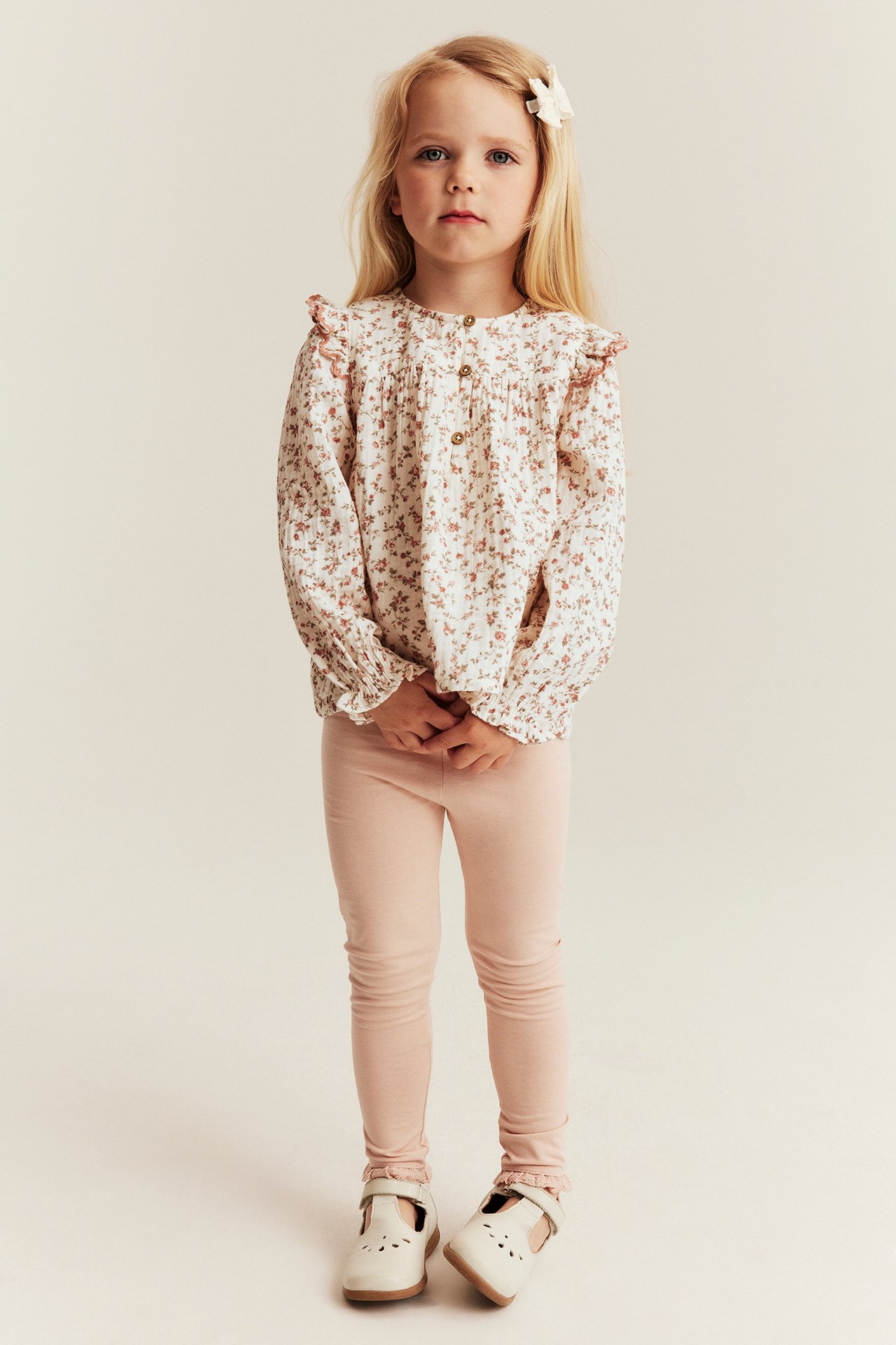 Front view: Kid in an off-white floral muslin blouse, long sleeves, pink leggings, and white shoes.