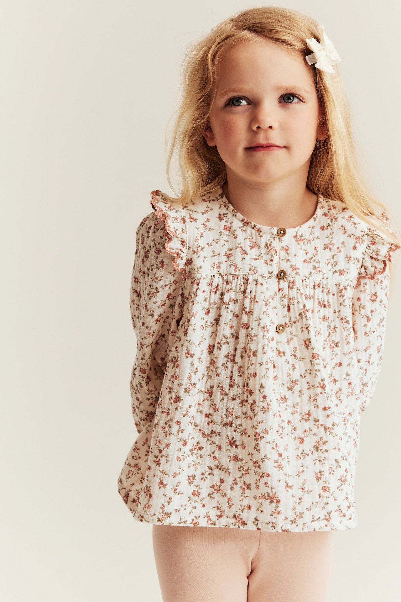 Vorderansicht: Kind in off-weißer Musselinbluse mit Blumenmuster und Schulterrüschen, dazu hellrosa Hose.