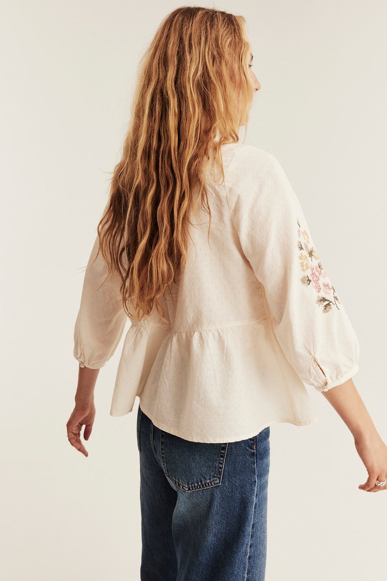 Rückenansicht einer Frau in einer cremefarbenen Peplum-Bluse mit floralen 3/4-Ärmeln und blauen Jeans.