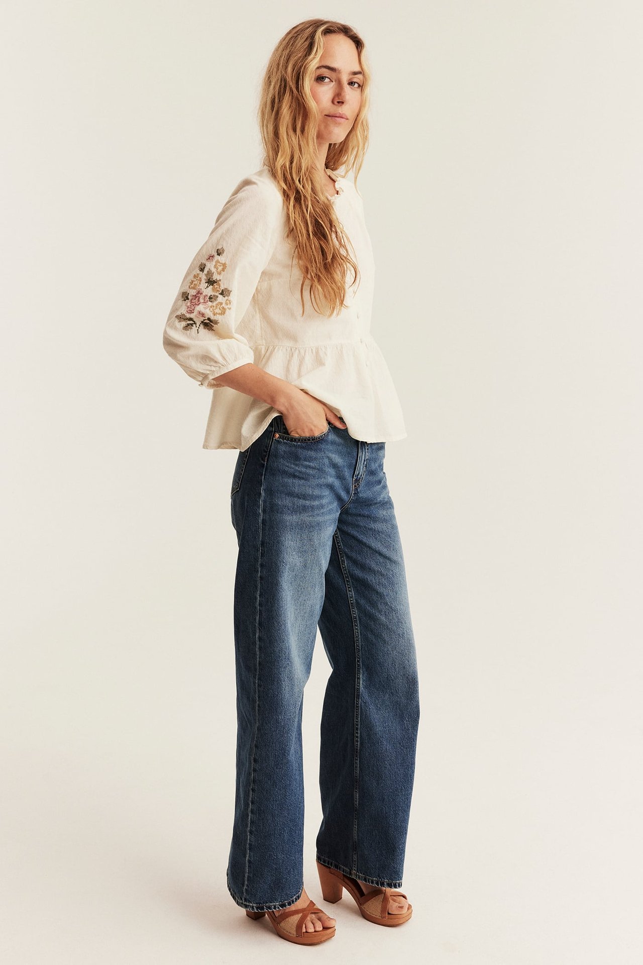 Vorderansicht einer Frau im cremefarbenen bestickten Peplum-Top, blauen Jeans und braunen Sandalen.
