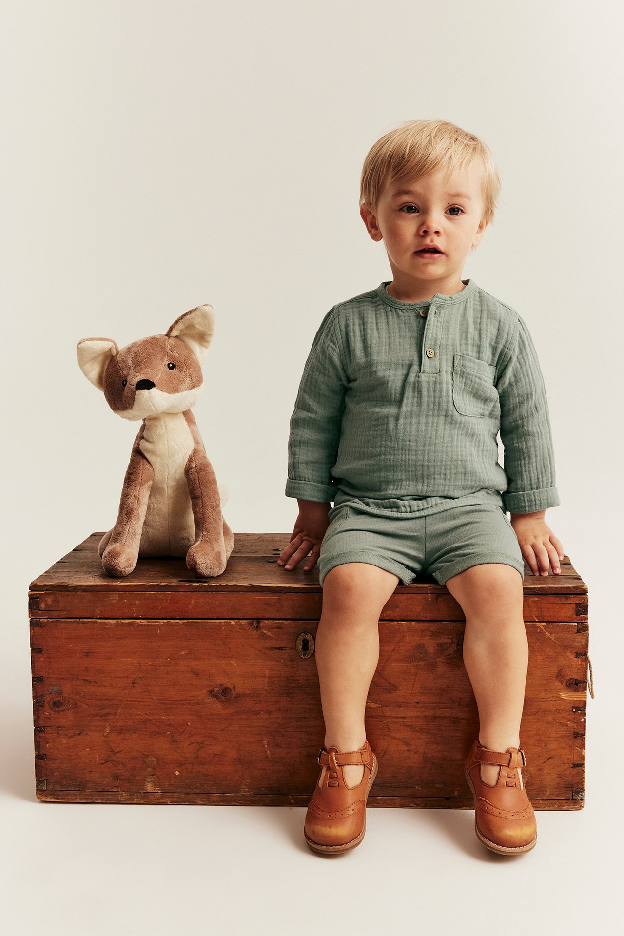 Vorderansicht: Kind in salbeigrünem Musselin-Set, braunen T-Bar-Schuhen, neben einem Fuchs.