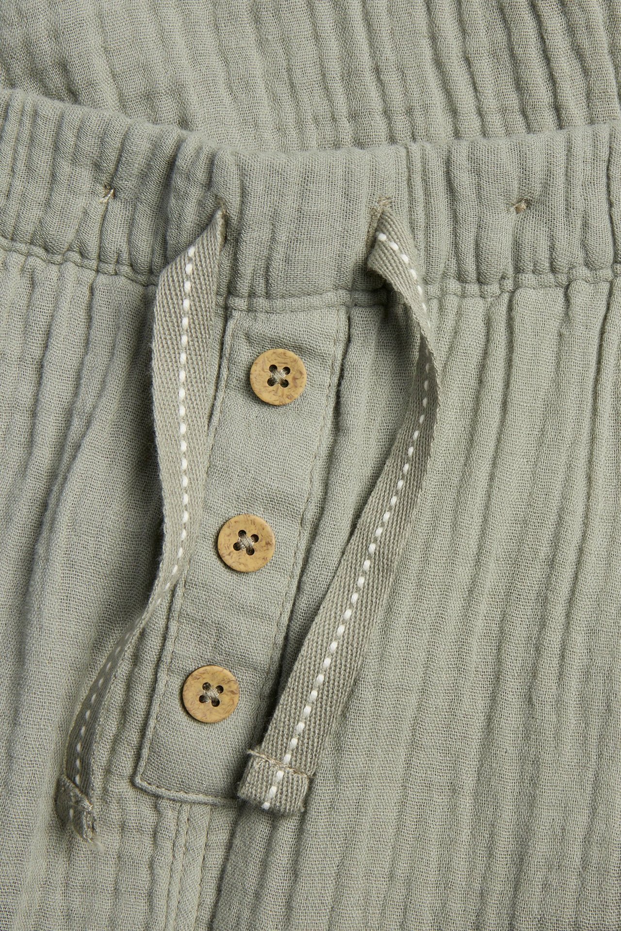 Nahaufnahme einer dunkelgrünen Baumwoll-Musselin-Hose mit Gummibund, Fischgrät-Kordelzug und dekorativen Holzknöpfen.