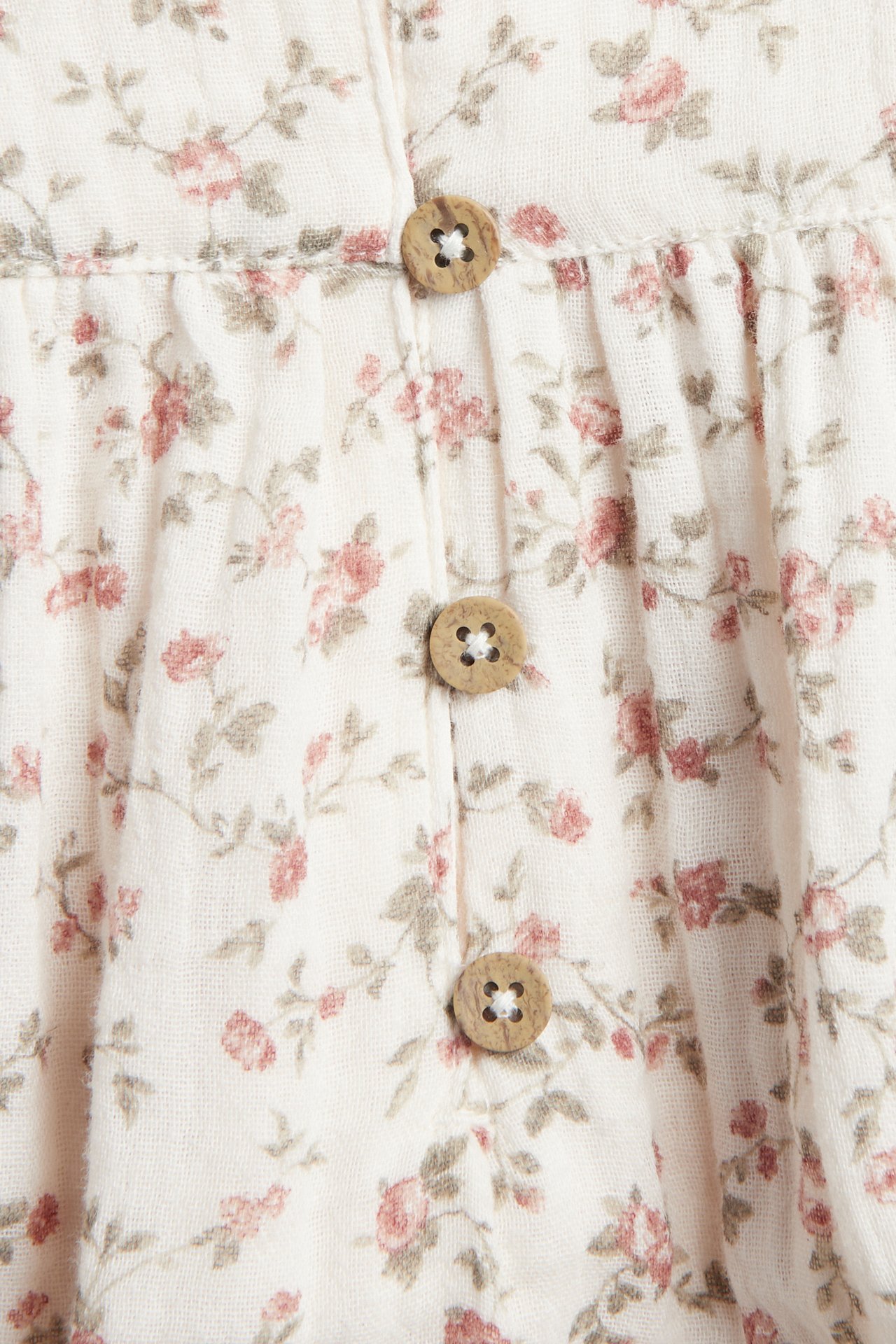 Nahaufnahme eines cremefarbenen Baby-Bodys aus Baumwollmusselin mit Blumenmuster und drei Holzknöpfen.