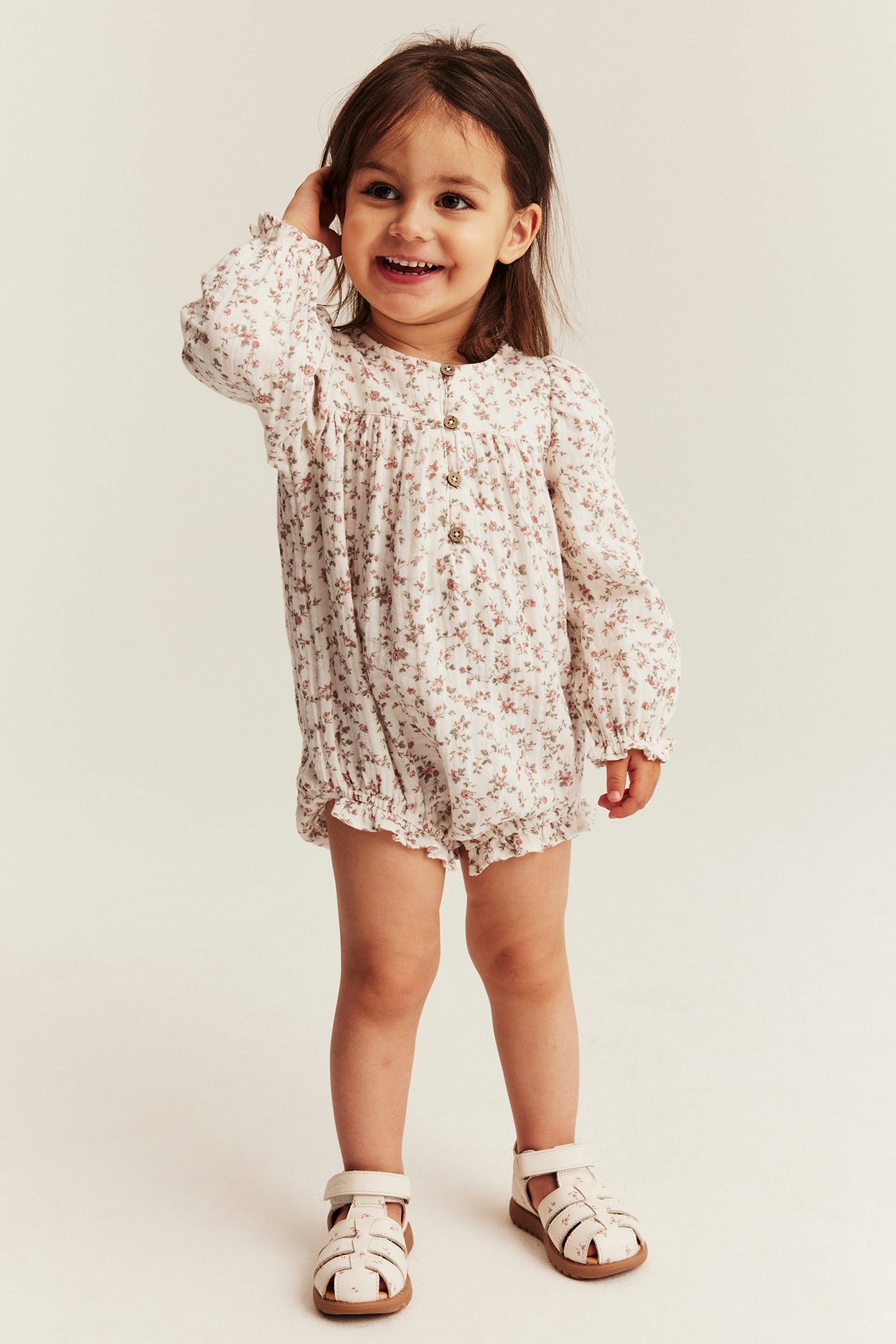 Front view of a smiling kid in an off-white floral muslin body with long sleeves and light beige sandals.