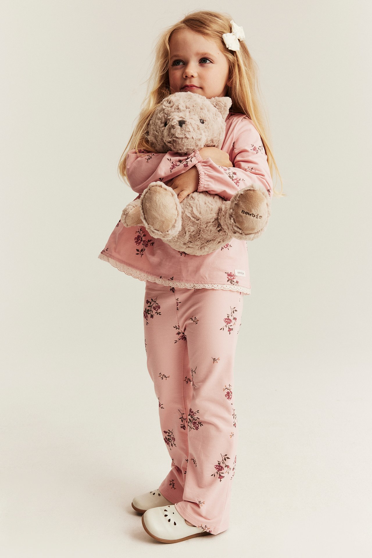 Front view: Kid wears a light pink floral cotton peplum top, matching pants, and holds a teddy bear.