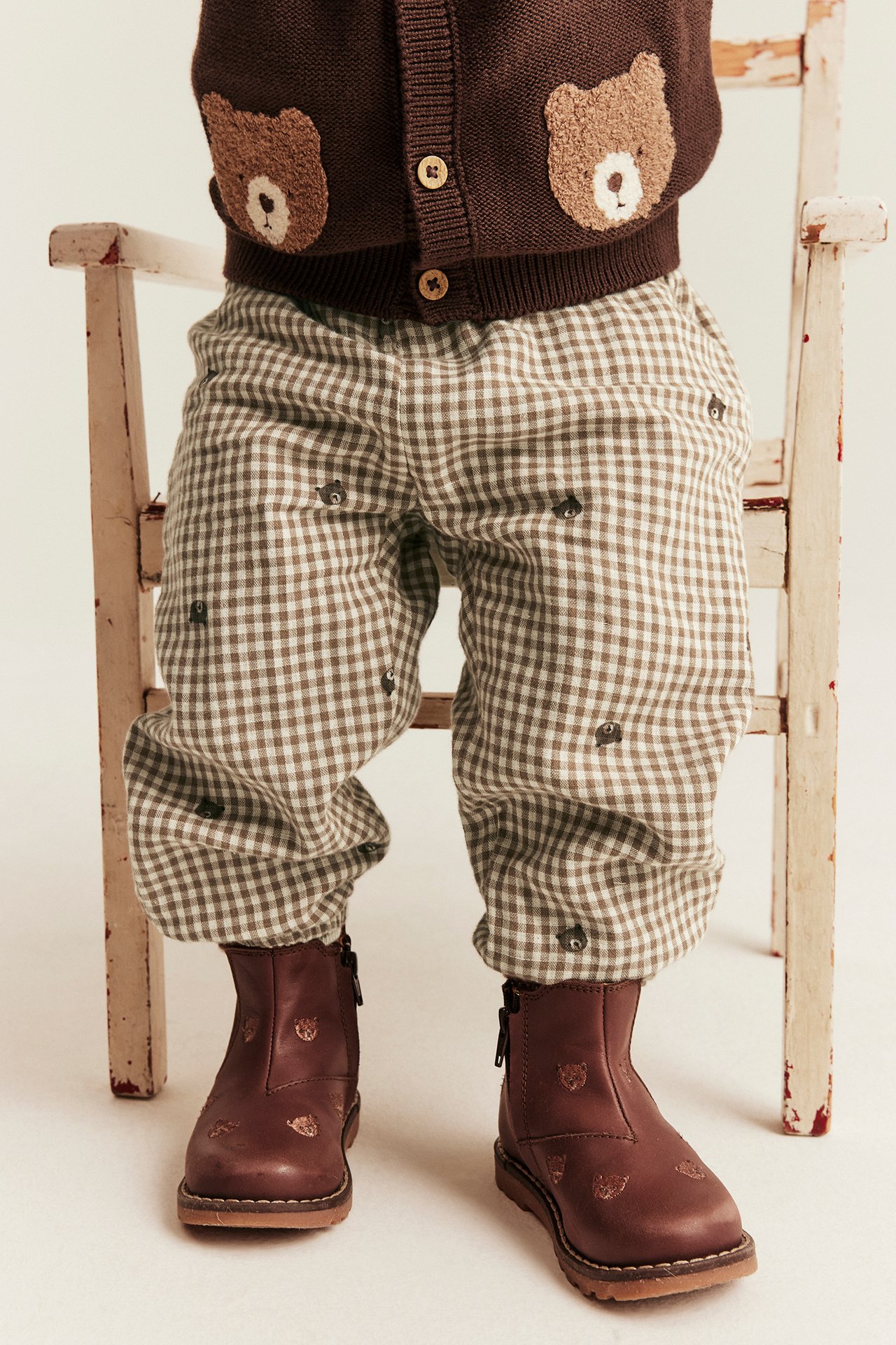 Vorderansicht: Kinder-Braun-Bärenstrickjacke, braune Vichy-Bärenhose, braune Lederstiefel.