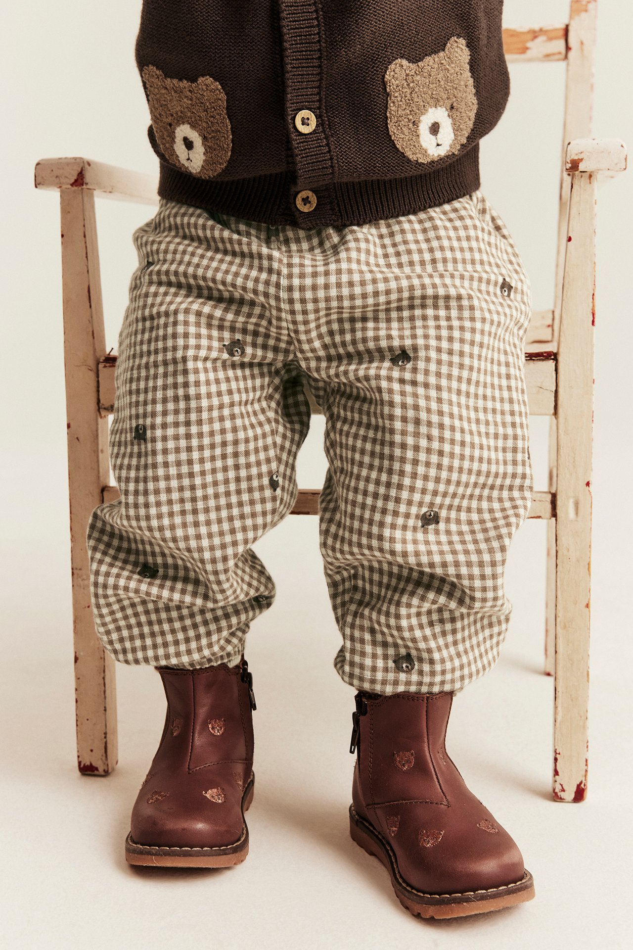 Front view of a kid's brown and white checkered pants, a brown bear applique cardigan, and brown boots.