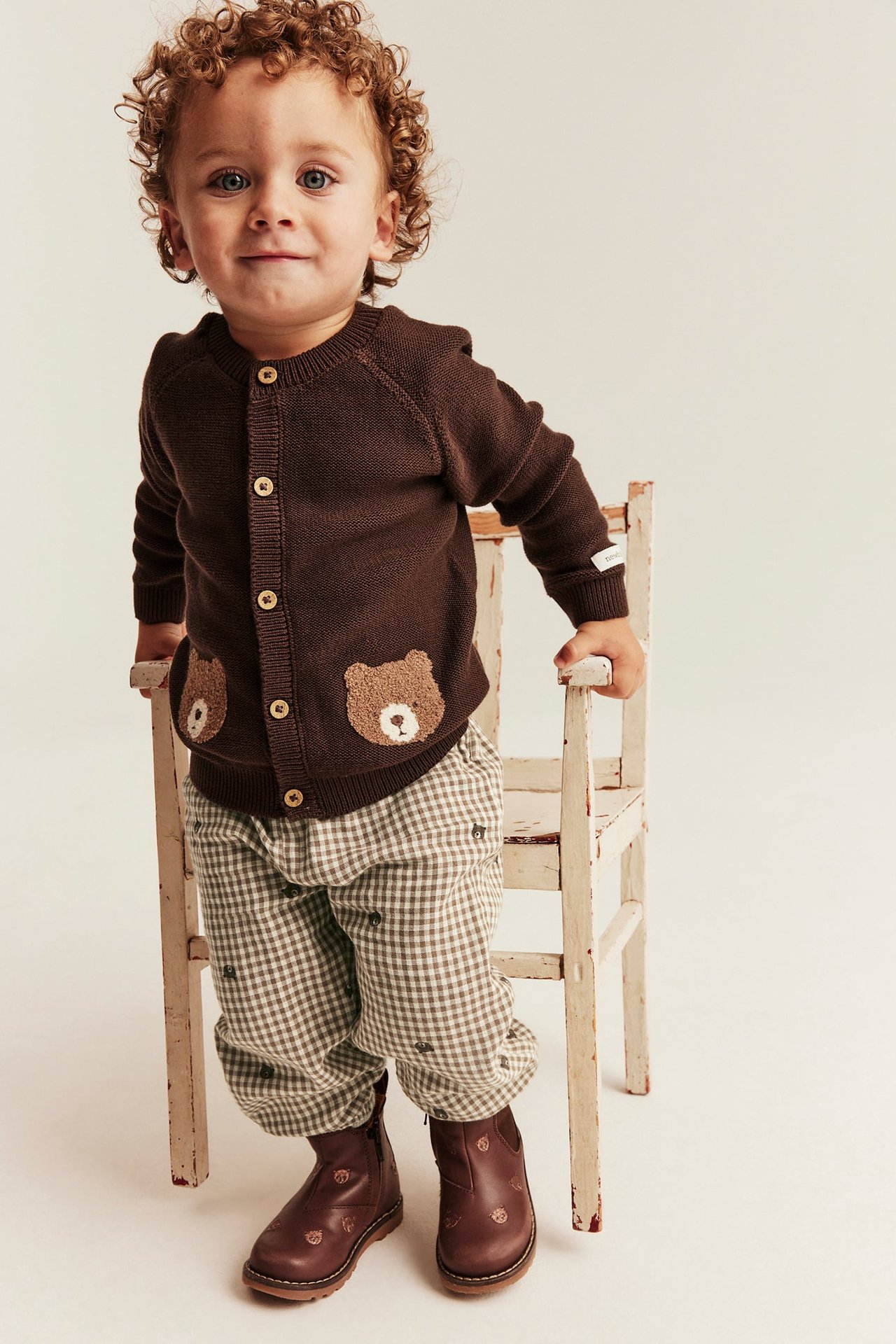Vorderansicht: Baby in brauner Strickjacke mit Bärenapplikationen, karierter Hose und braunen Stiefeln.
