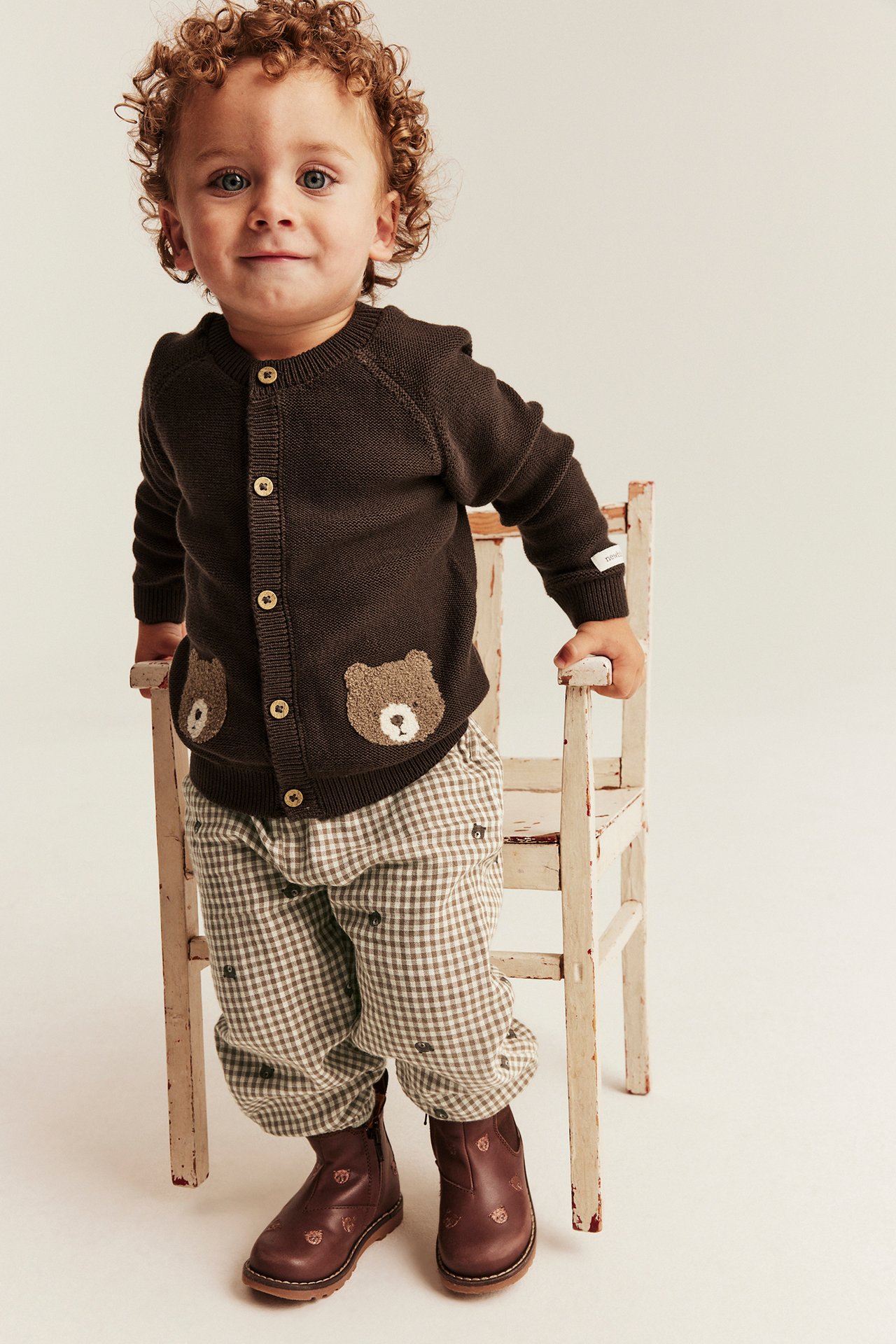 Front view of a baby in a brown cardigan with bear pockets, gingham pants, and brown boots.