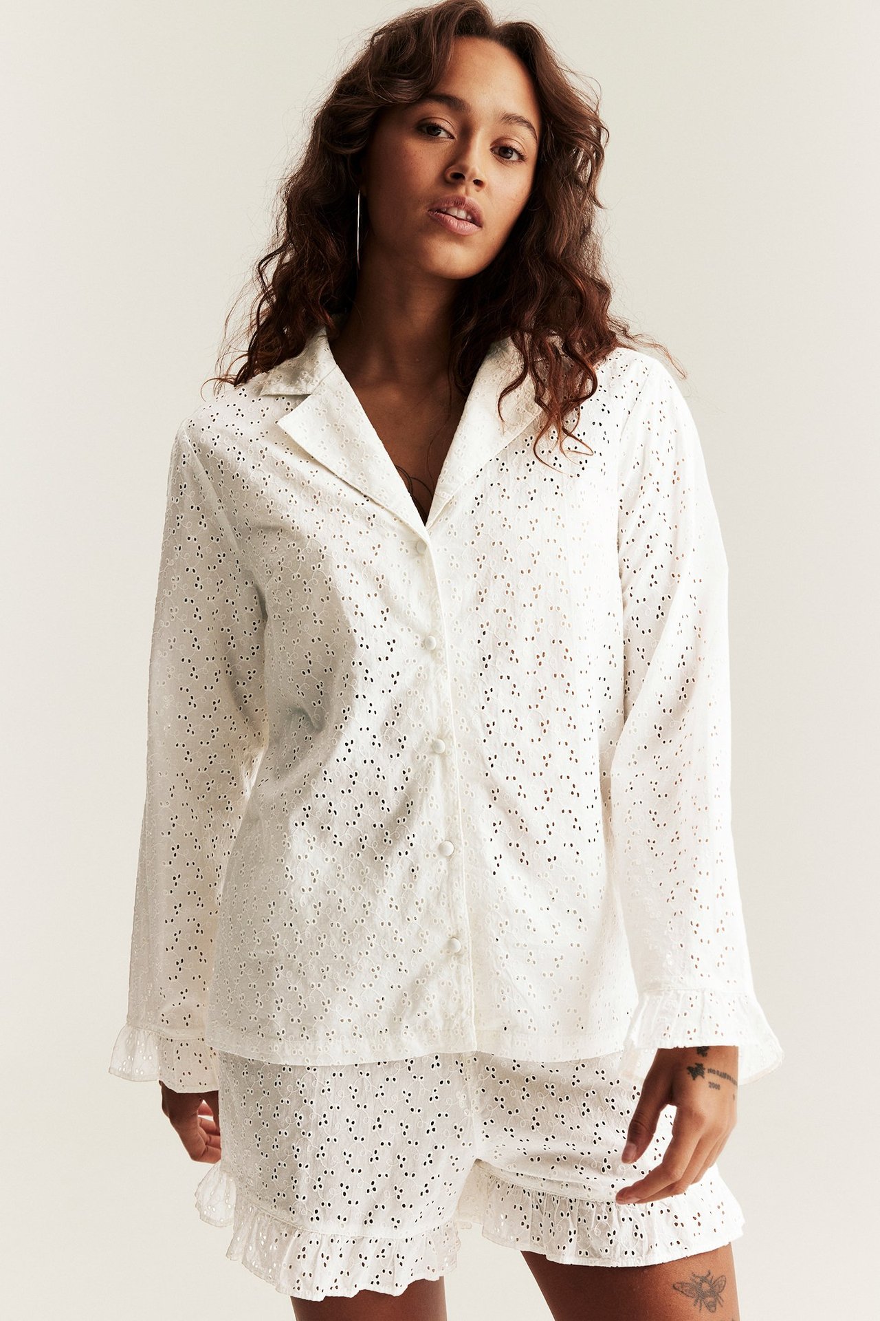 Front view of a woman wearing an off-white cotton broderie anglaise pyjama set with ruffles.
