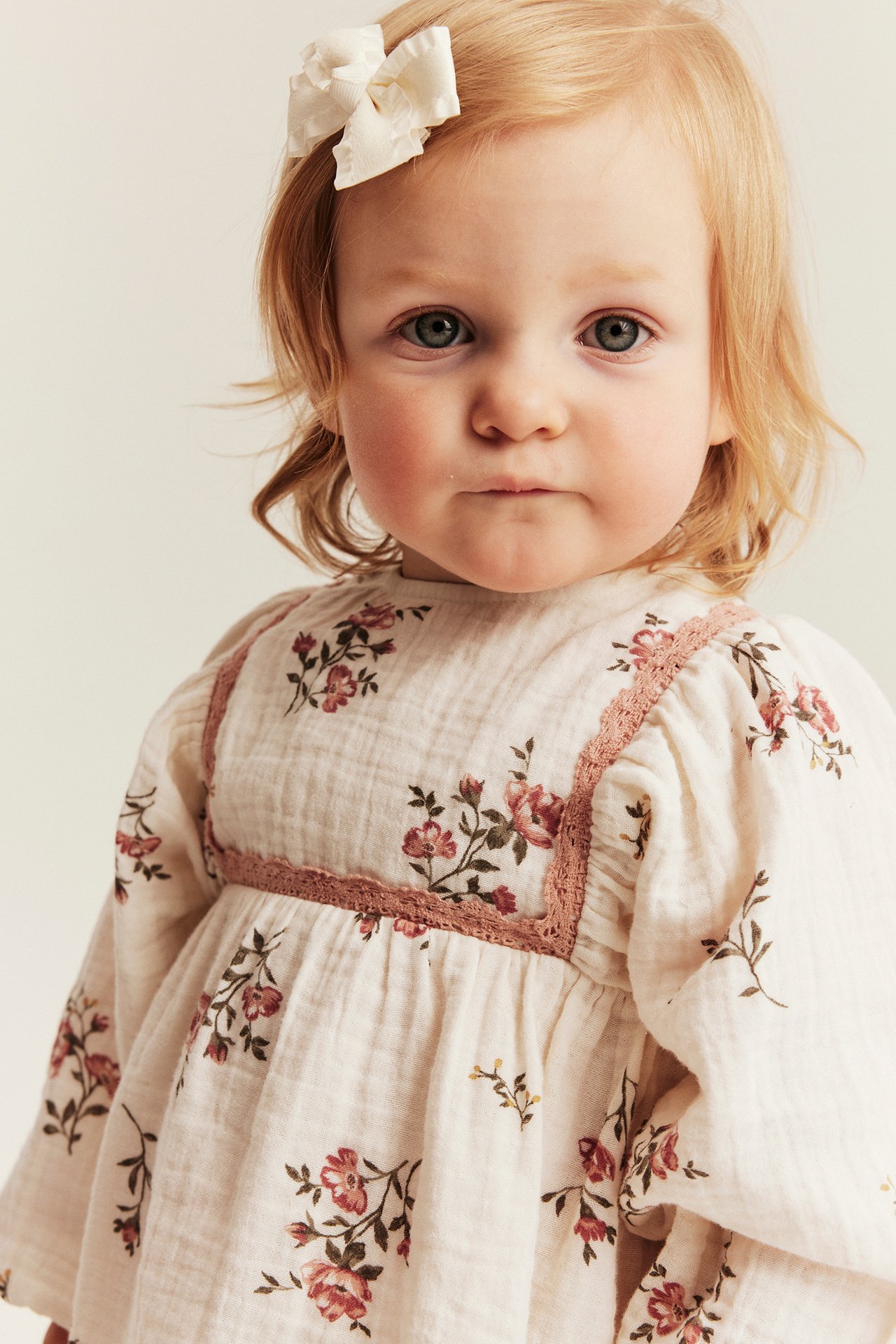 Close-up of a baby wearing an offwhite floral muslin dress with pink lace and an offwhite hair bow.
