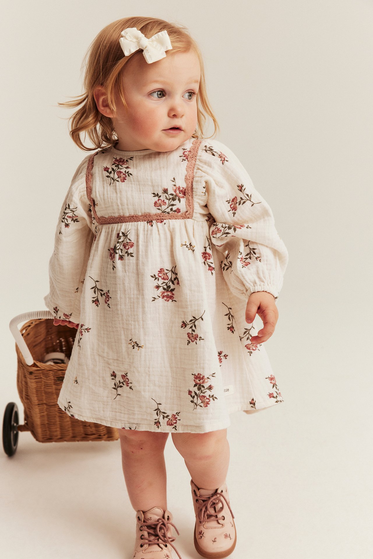 Front view: Baby girl in an off-white floral muslin dress with pink lace, long sleeves, and pink boots.