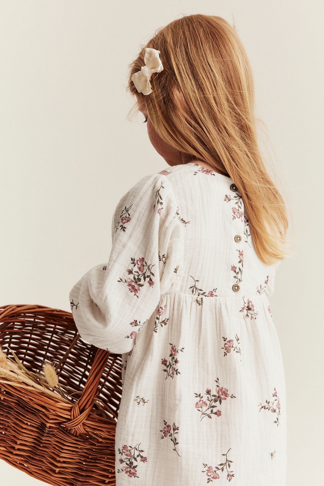 Back view: Kid in offwhite floral muslin dress with back buttons. Wears bow, holds wicker basket.