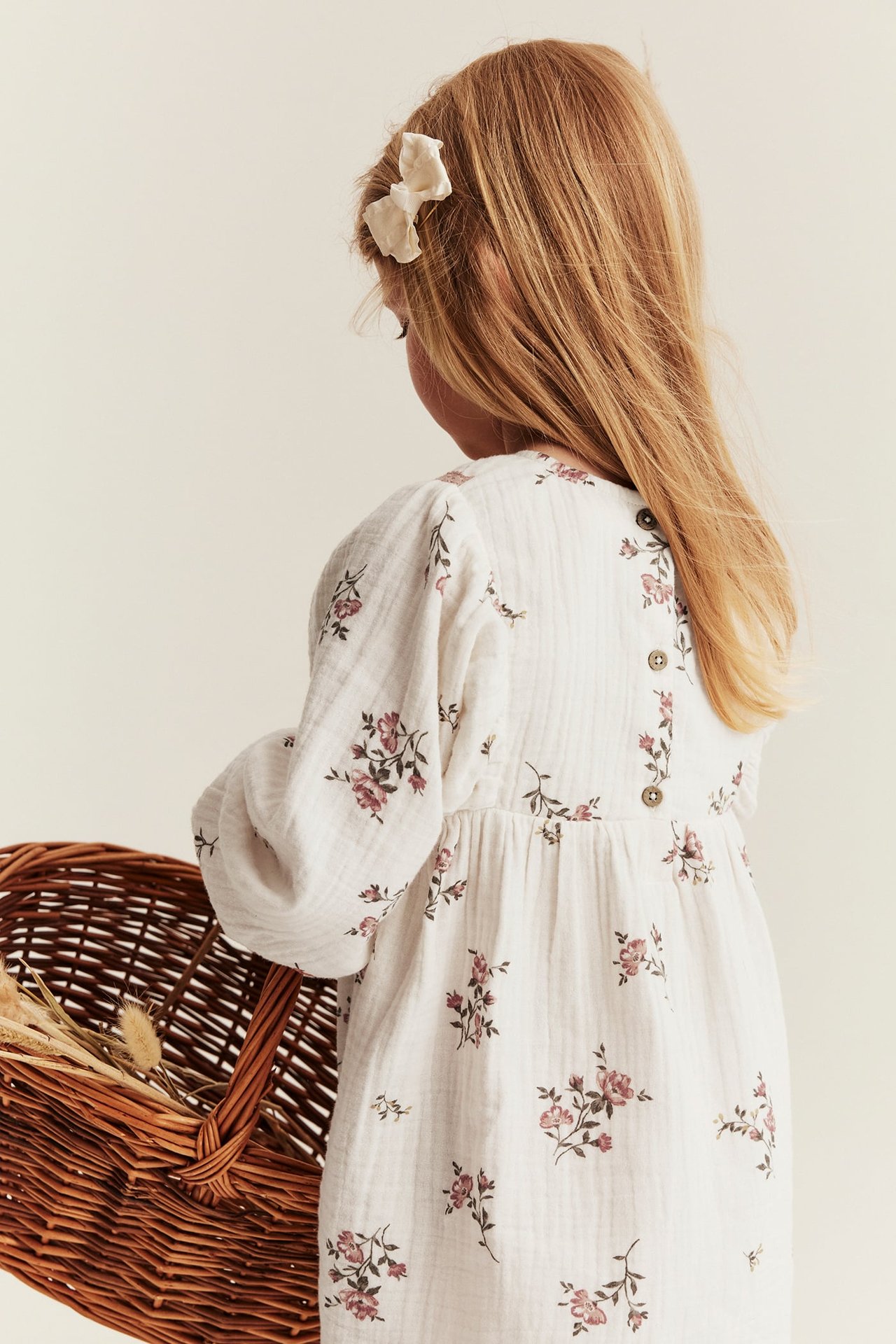 Rückenansicht: Kind in offwhite geblümtem Musselin-Kleid mit Rückenknöpfen. Trägt Schleife, hält Weidenkorb.