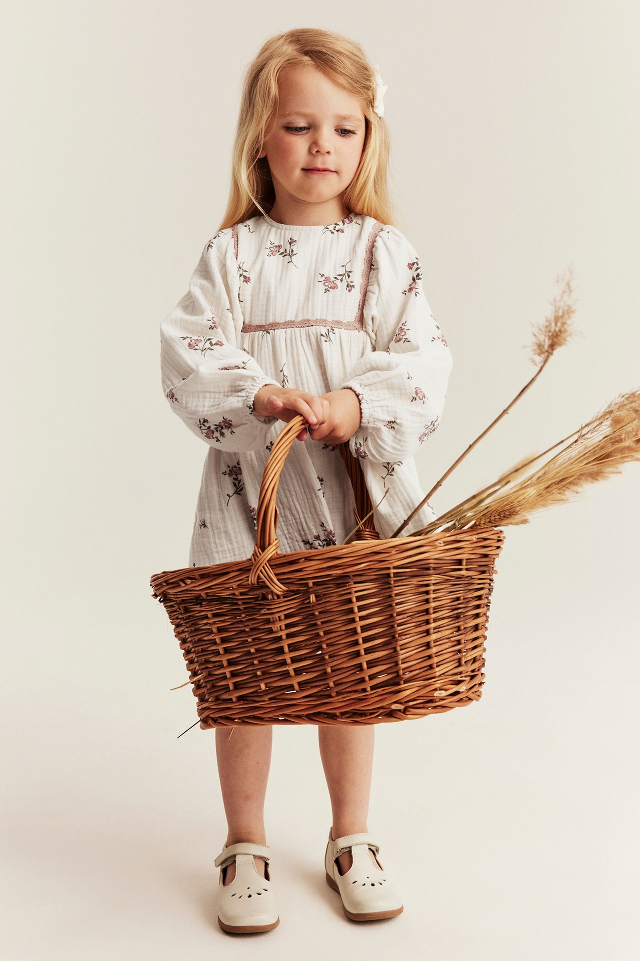 Front view: Kid in offwhite floral muslin dress with lace trim, holding a wicker basket, light beige shoes.