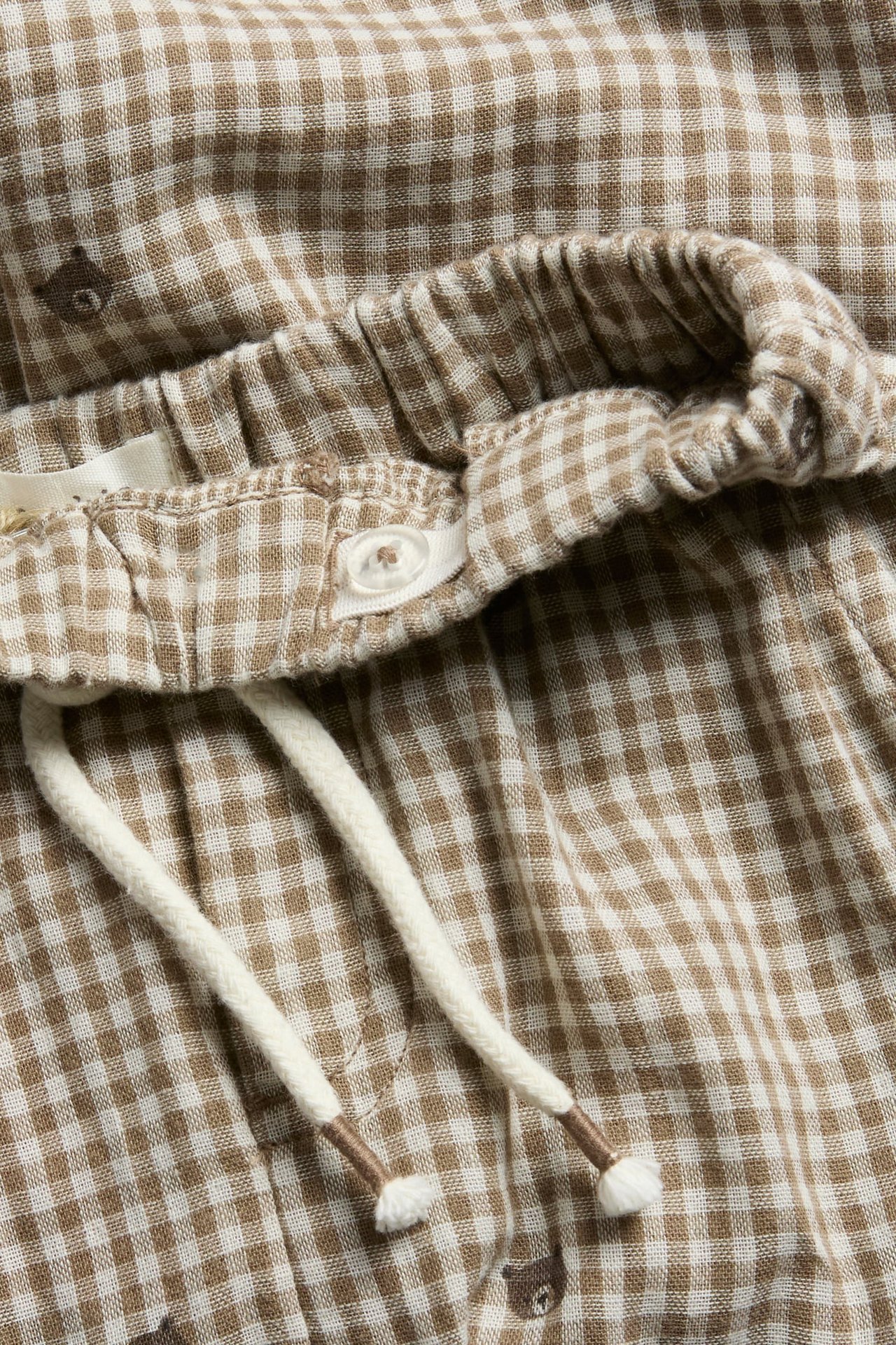 Nahaufnahme von braun-weißen Vichy-Shorts mit Gummizug, Knopf, Kordelzug und Bärendruck.