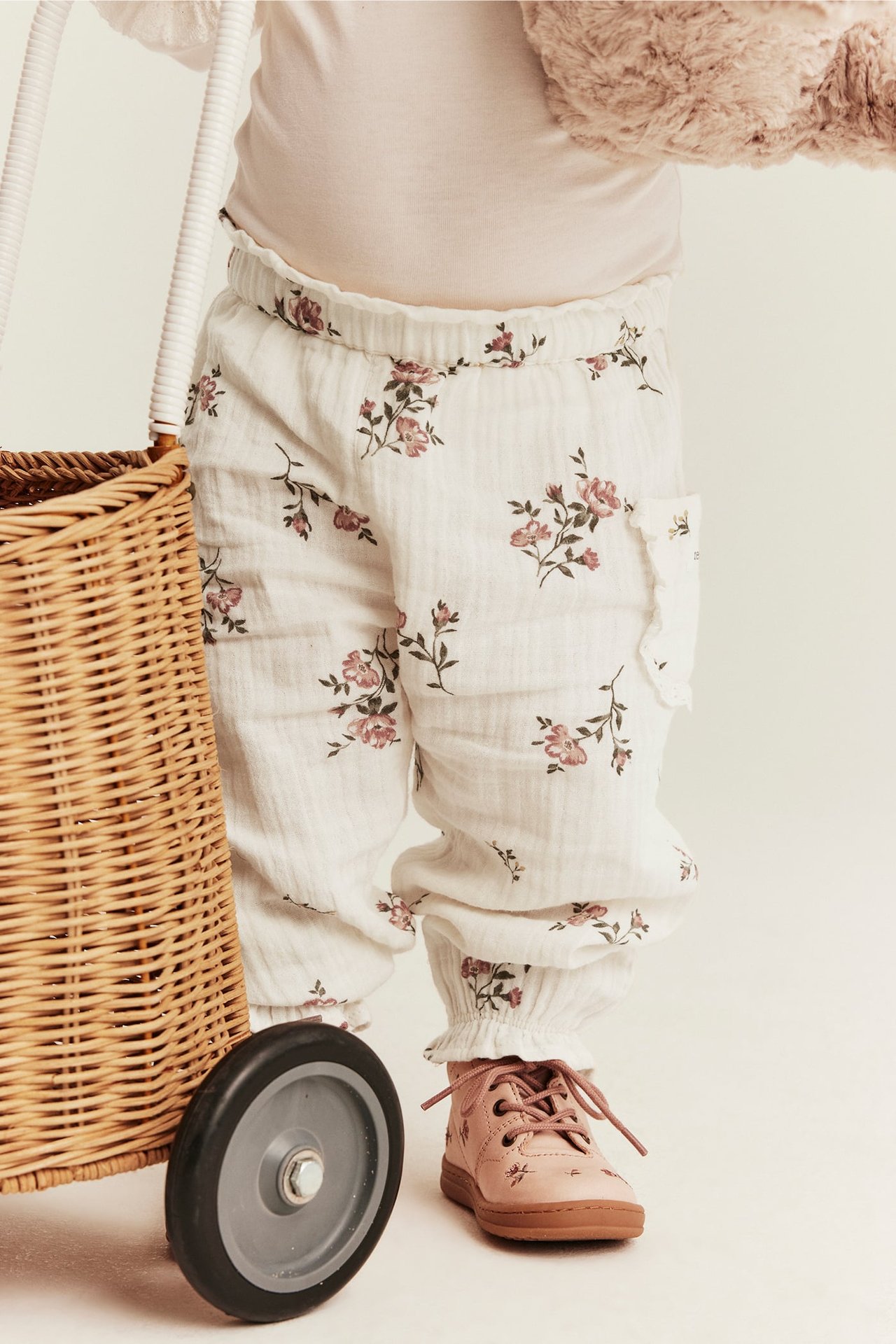 Vorderansicht: Baby trägt cremeweiße florale Baumwoll-Puffhose mit Spitzentaschen und rosa Schnürschuhe.