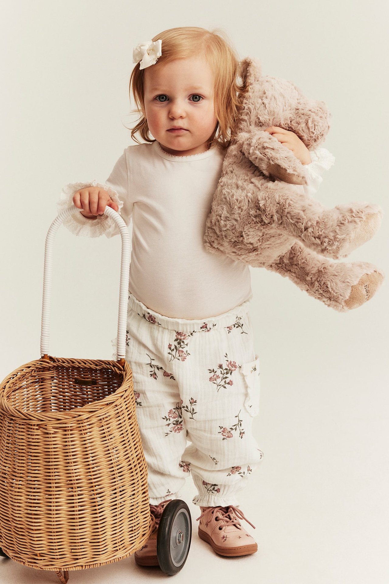 Vorderansicht: Baby trägt off-white geblümte Webhose, weißes Oberteil, rosa Schuhe, hält Teddy und schiebt Korbtrolley.