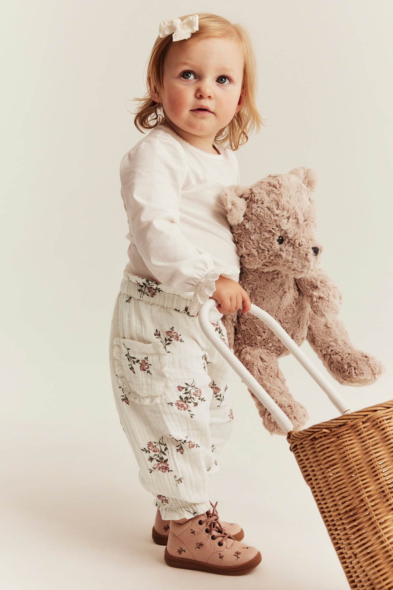 Vorderansicht Baby in cremeweißer geblümter Pumphose mit Spitzentaschen, weißem Oberteil, pinken Stiefeln, hält Teddybär und Korb.