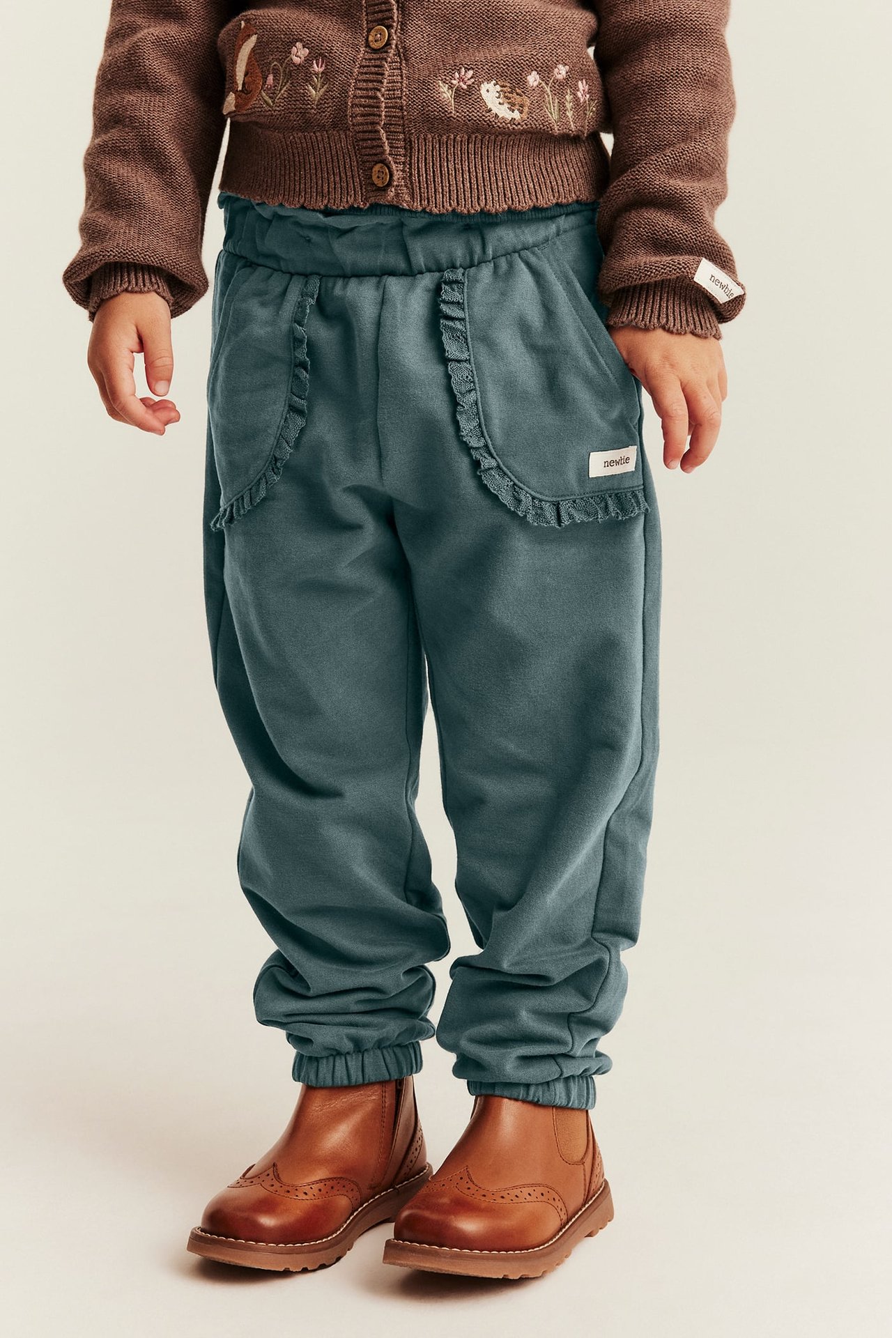 Front view of kid in dark teal sweatpants with ruffled pockets, brown knitted cardigan and brown boots.