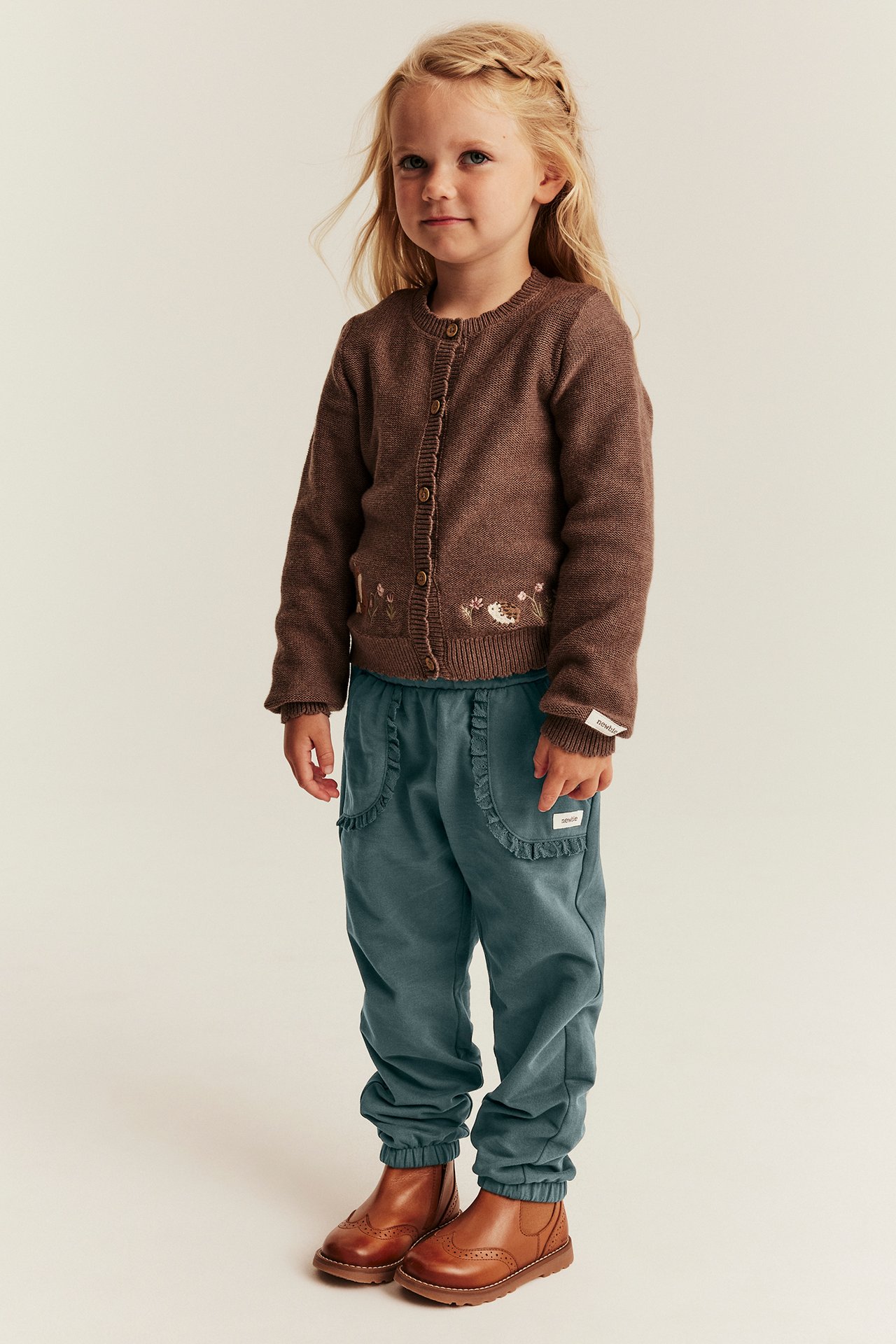 Vorderansicht: Kind in brauner bestickter Strickjacke, teal Rüschen-Sweatpants, braunen Lederstiefeln.