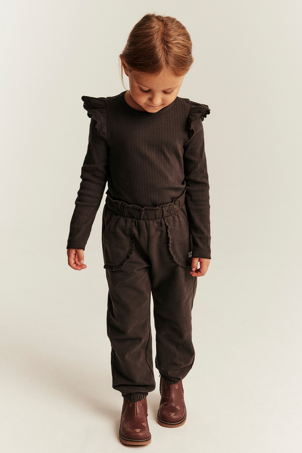 Kid in dark brown ribbed long-sleeve top with ruffles, matching sweatpants, and brown embroidered boots.