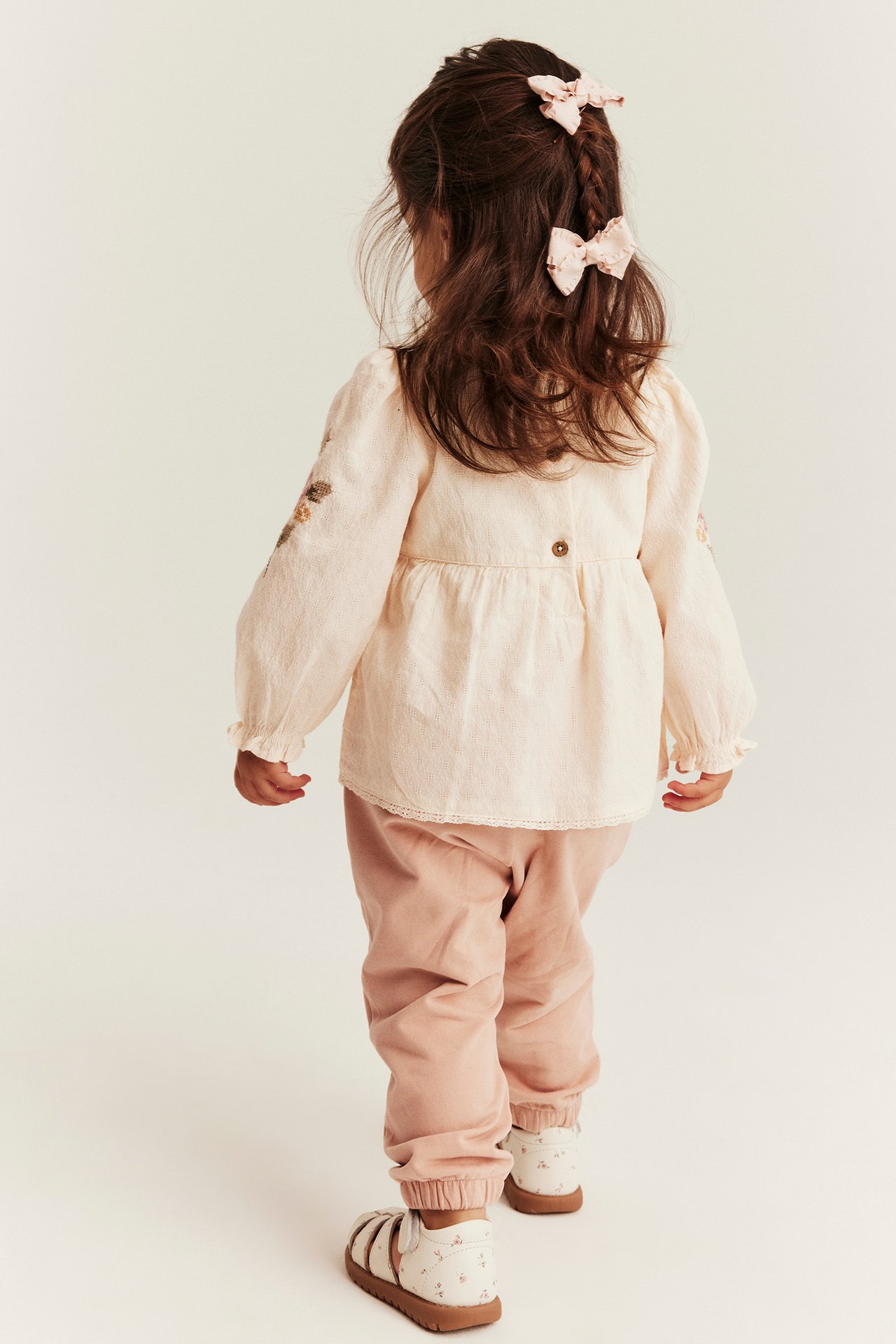 Kid in off-white embroidered peplum bodysuit with back buttons, pink pants, floral sandals, two pink bows.