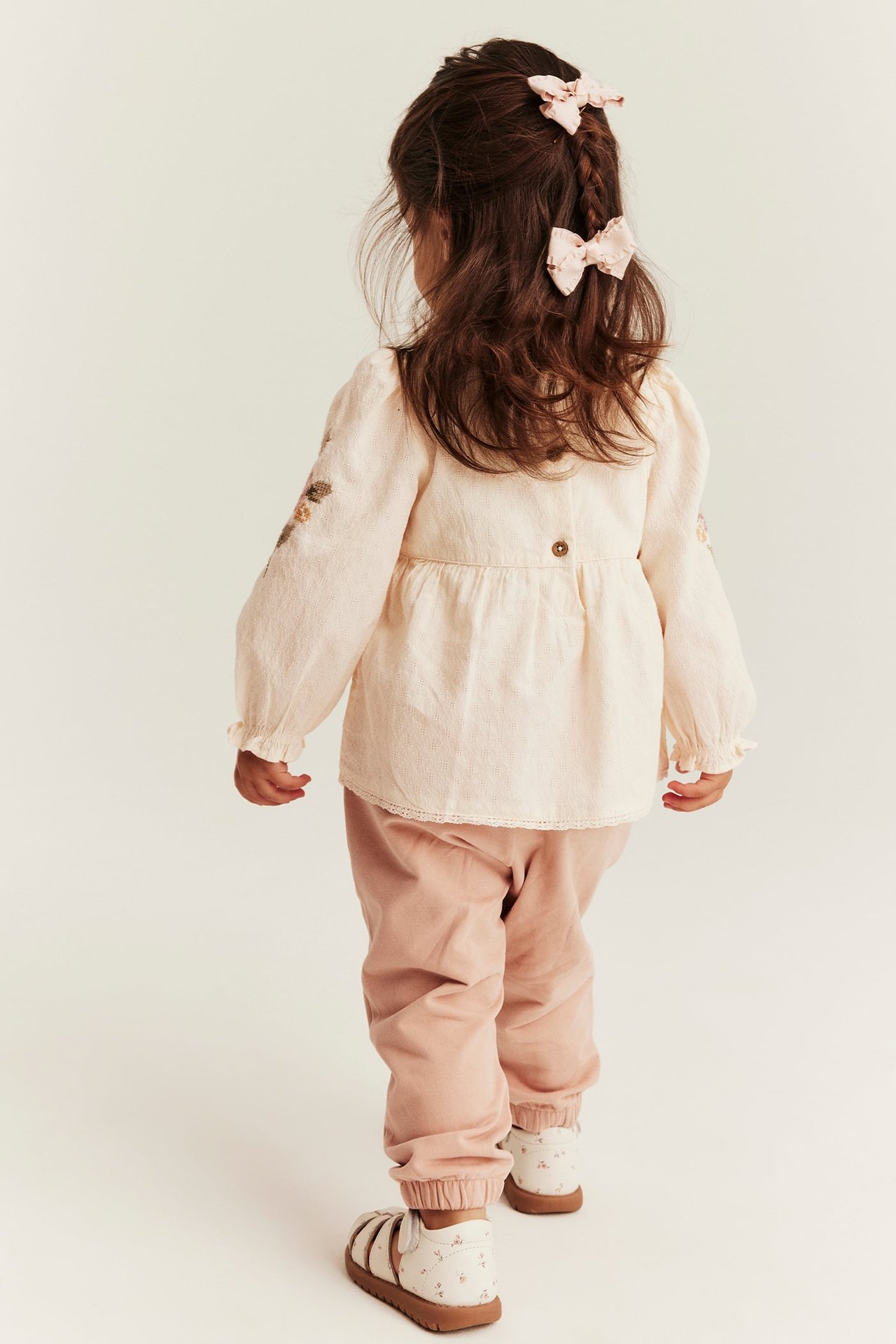 Kind in cremefarbenem bestickten Peplum-Body mit Rückenknöpfen, rosa Hose, geblümten Sandalen und zwei rosa Schleifen.