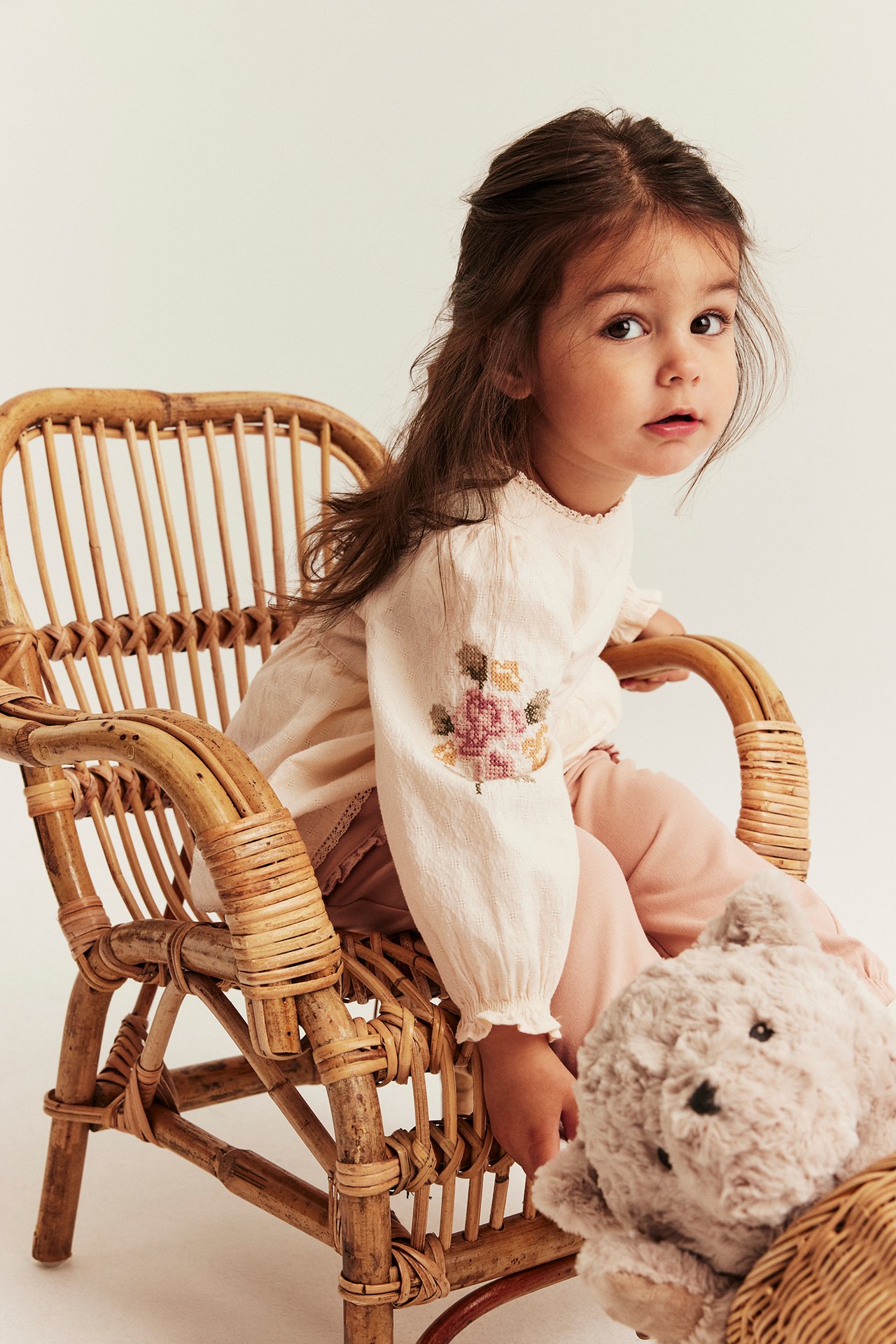 Front view: Kid in off-white embroidered woven bodysuit, pink trousers, with a teddy bear.