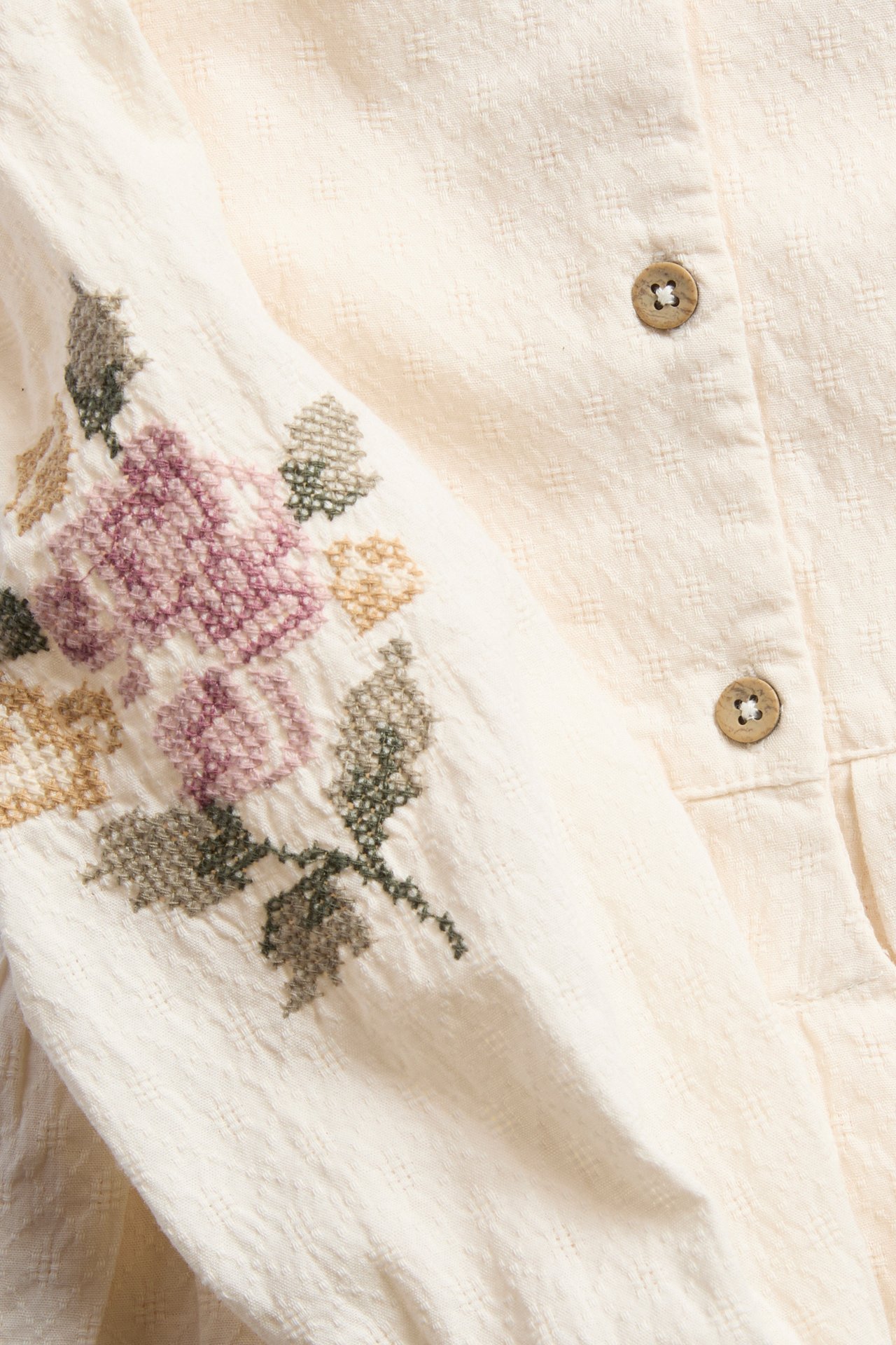 Close-up: offwhite textured woven baby bodysuit with cross-stitch flower embroidery on sleeve, back buttons.