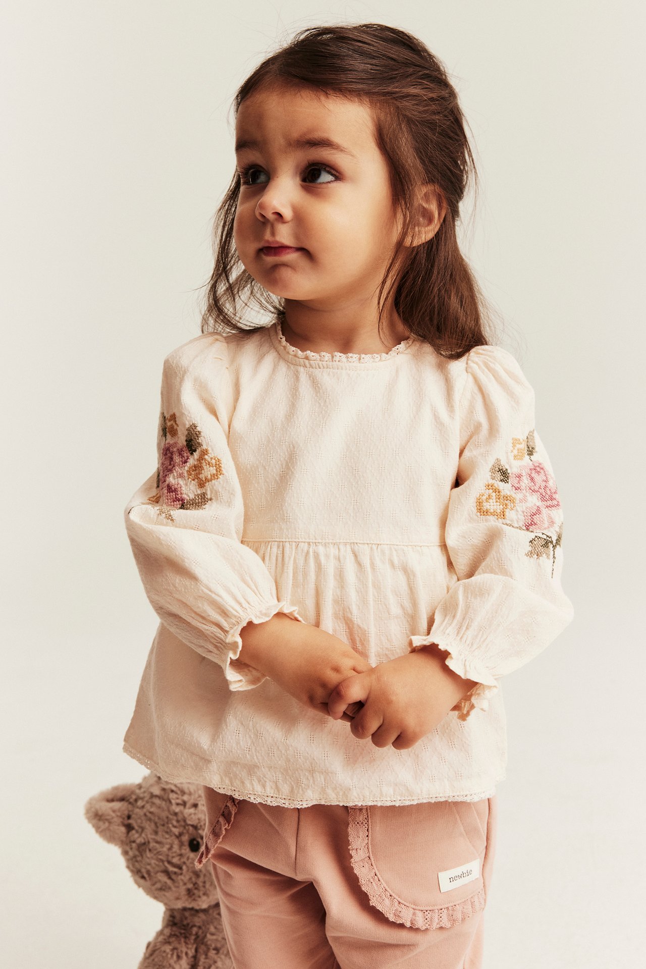 Front view of a kid wearing an off-white woven peplum blouse-body with floral embroidery, and pink pants.