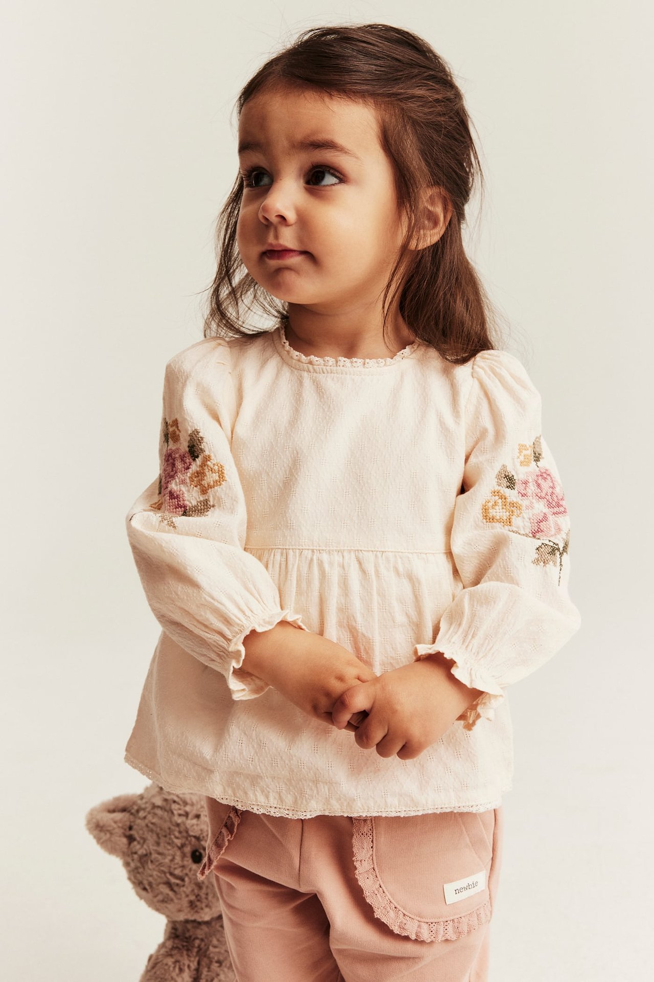 Vorderansicht: Kind trägt einen off-weißen gewebten Peplum-Blusenbody mit Blumenstickerei und rosa Hose.