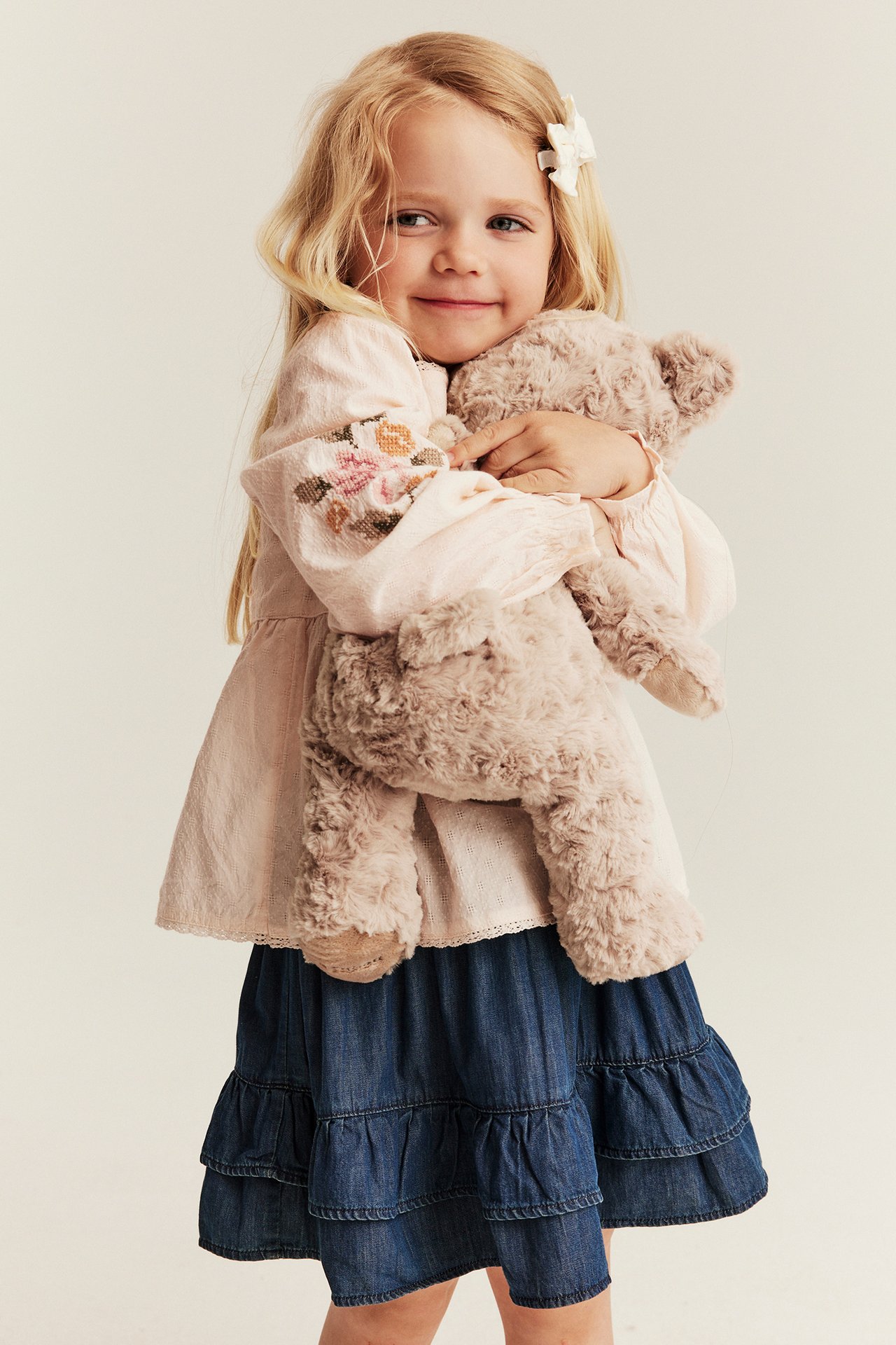 Front view: kid in off-white embroidered cotton blouse, blue tiered skirt, holding a beige teddy bear.