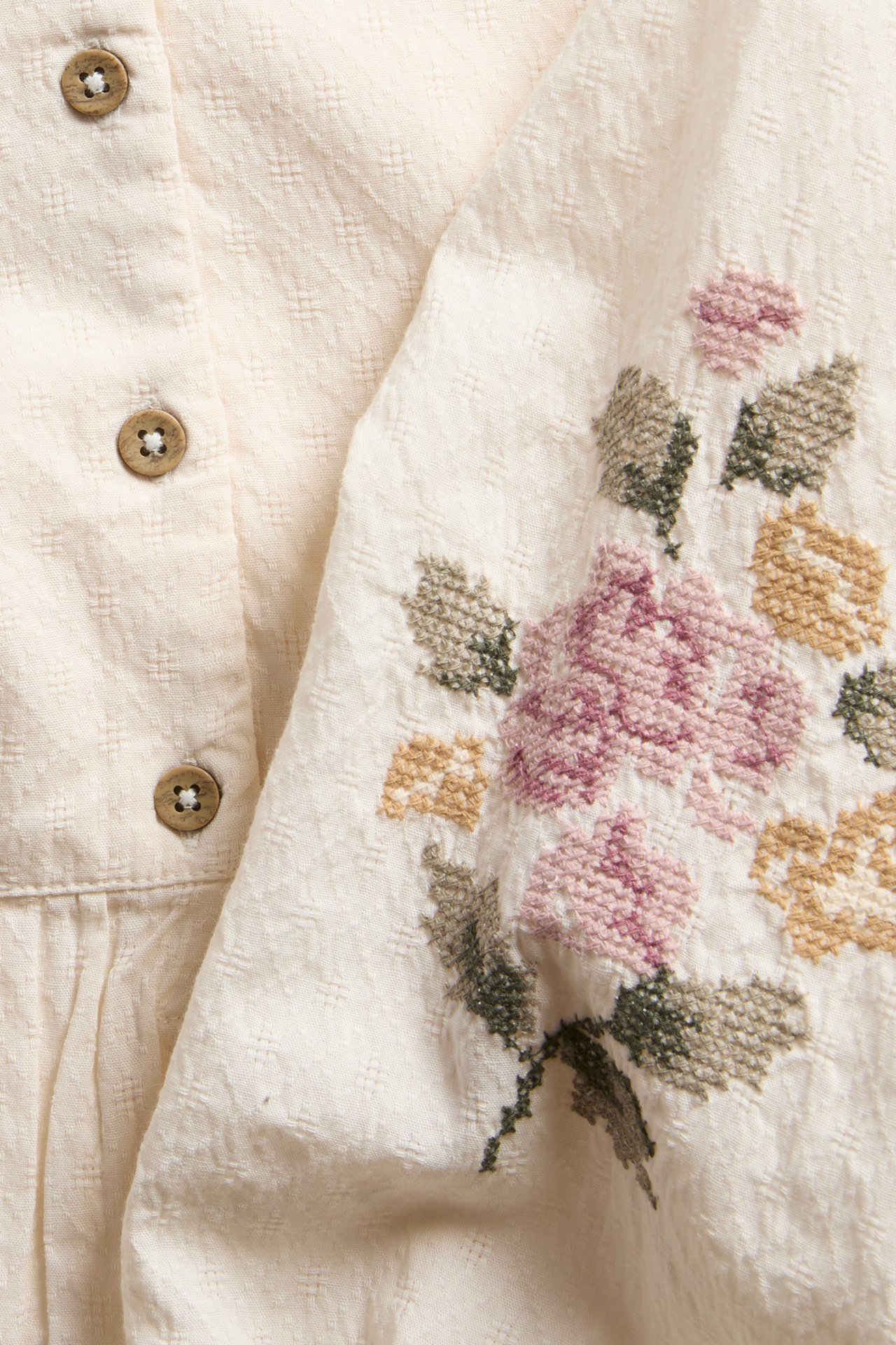 Close-up of an off-white woven cotton blouse with textured fabric, colorful cross-stitch floral embroidery, and wooden buttons.