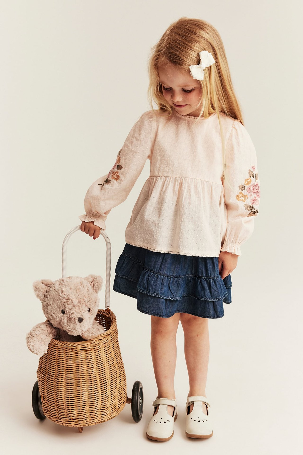 Vorderansicht: Kind in cremefarbener Baumwollbluse mit Blumenstickerei, blauem Jeansrock, weißen Schuhen, zieht Spielzeugwagen.