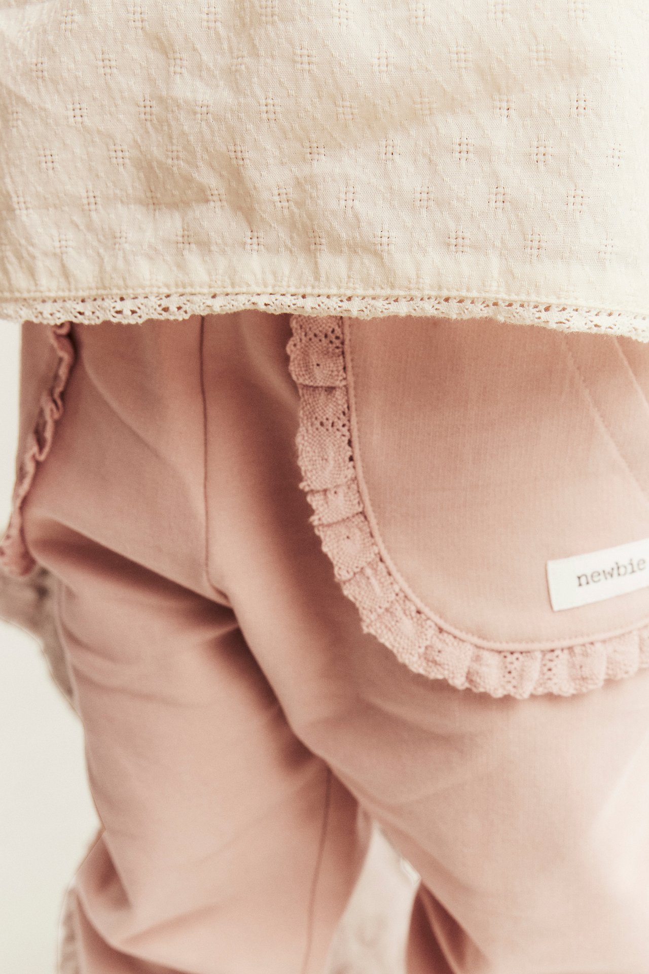 Nahaufnahme eines Babys in rosa Baumwoll-Jogginghose mit Spitzentaschen unter einem beigen gemusterten Oberteil.
