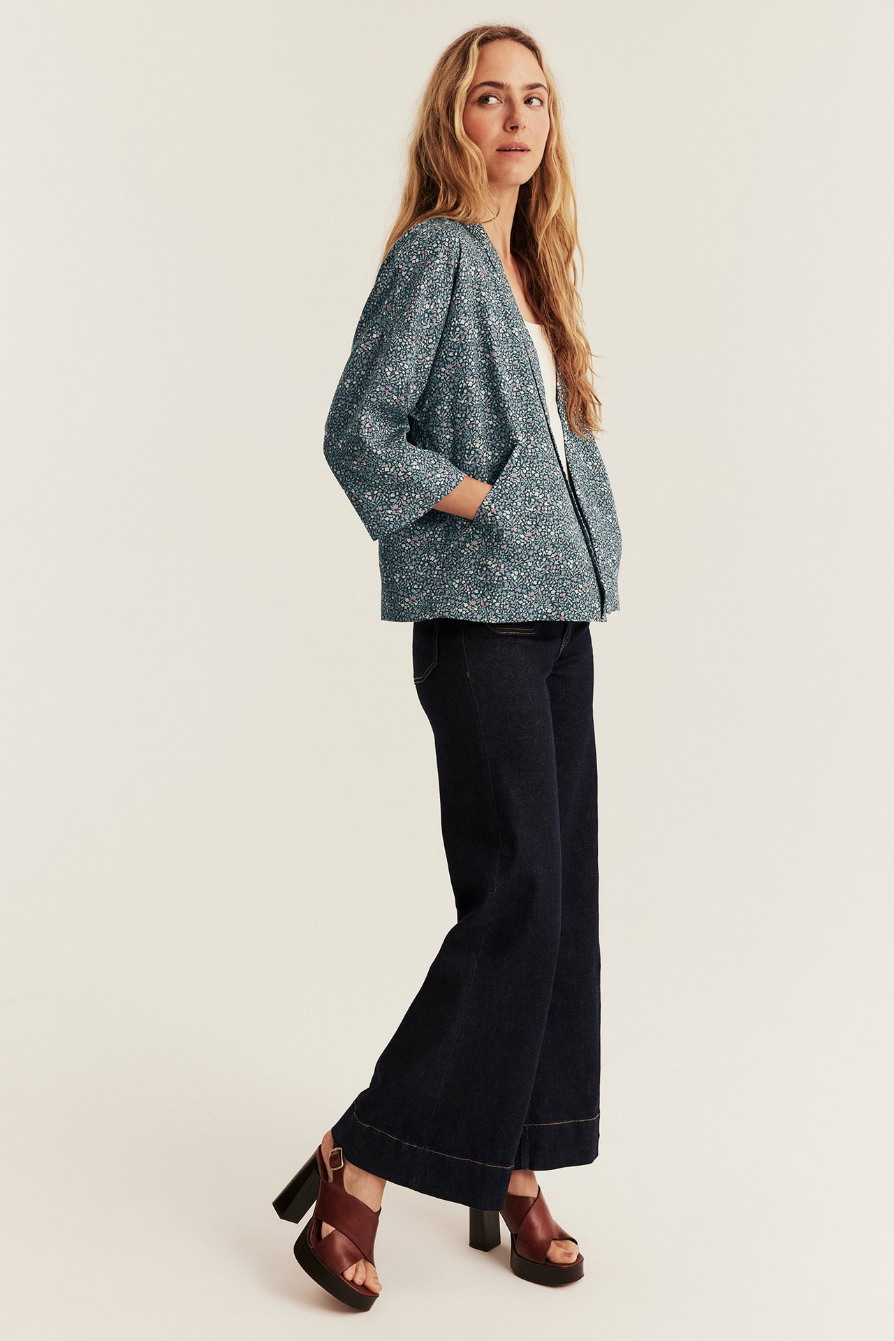 Seitenansicht einer Frau in einer teal-gemusterten Jacke, dunklen weiten Jeans und braunen Plateau-Sandalen.