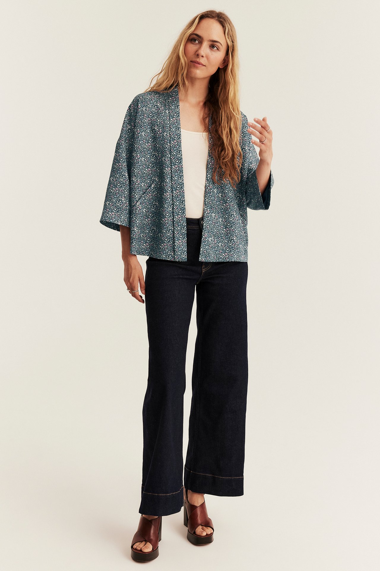 Frau in blauer floraler Kimonojacke, weißem Tanktop, dunklen Jeans und braunen Plateausandalen.